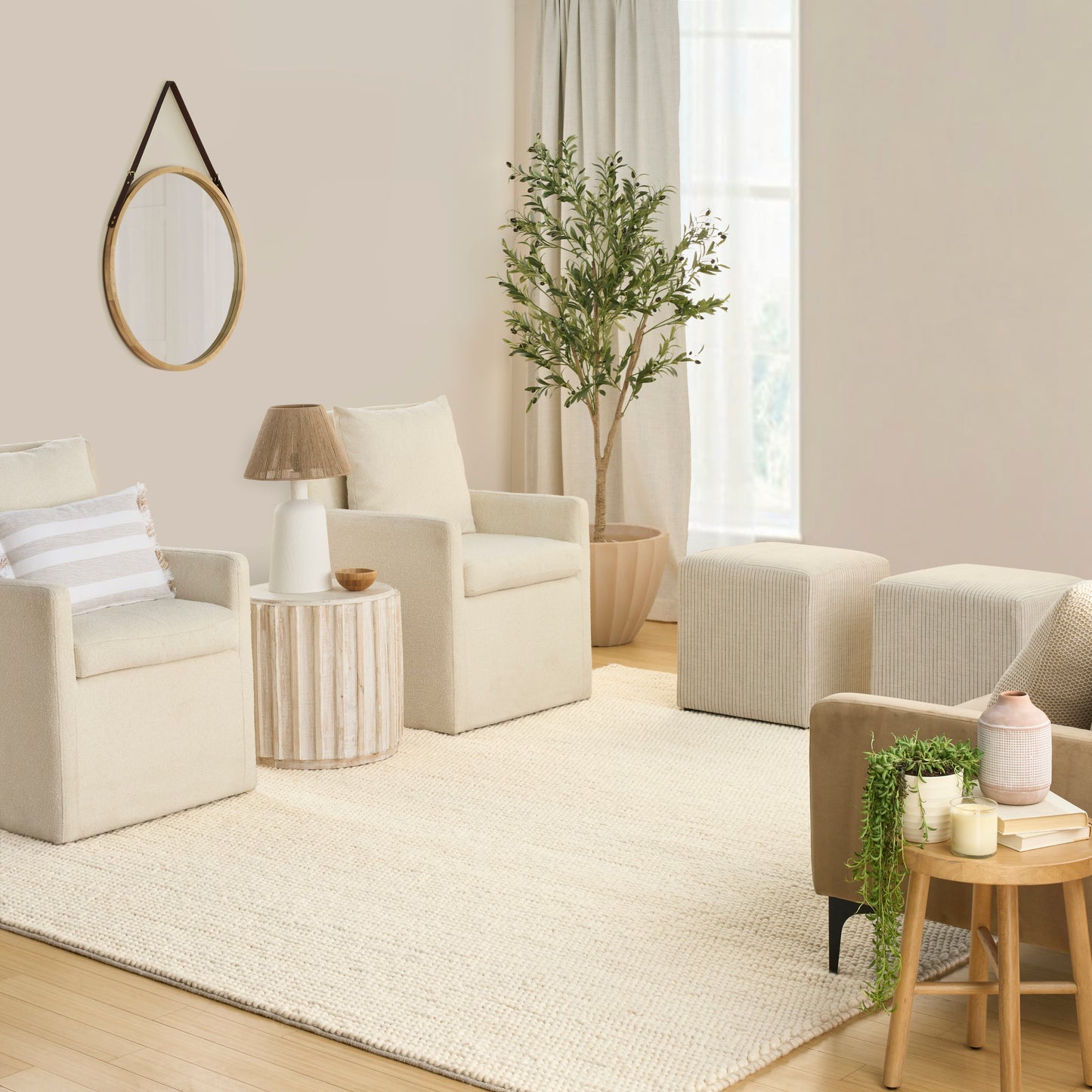 Neutral beige living room with two armchairs, matching ottomans, round wall mirror, tall plant, cream rug, light wood tables.