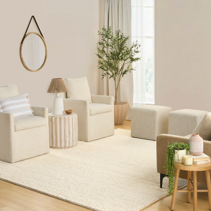 Neutral beige living room with two armchairs, matching ottomans, round wall mirror, tall plant, cream rug, light wood tables.