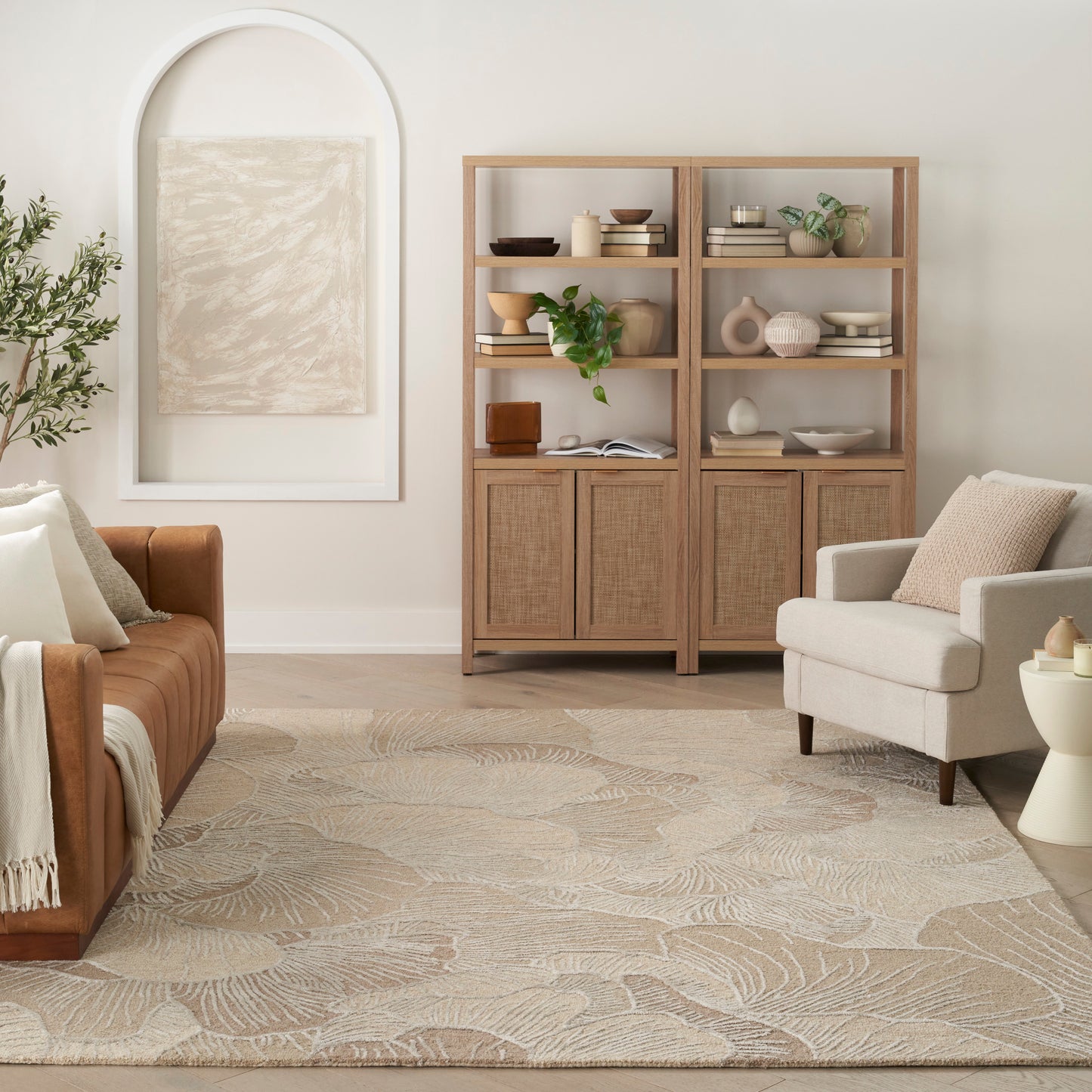 Light wood cabinet with glass doors and open shelves, beige armchair, brown leather sofa, and leaf-patterned neutral rug in a cozy living room.