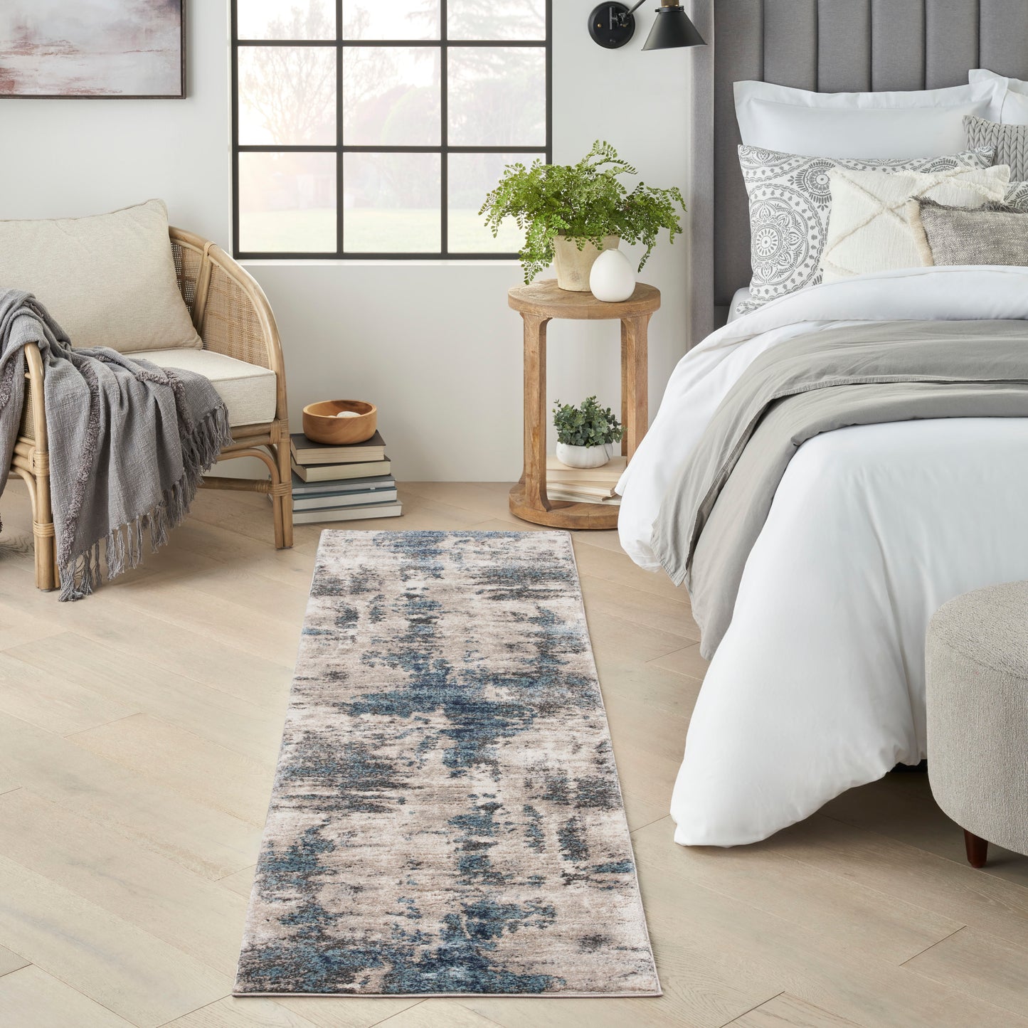 Abstract blue and beige runner rug along a light wood floor beside a white bed and rattan chair in a bright bedroom.