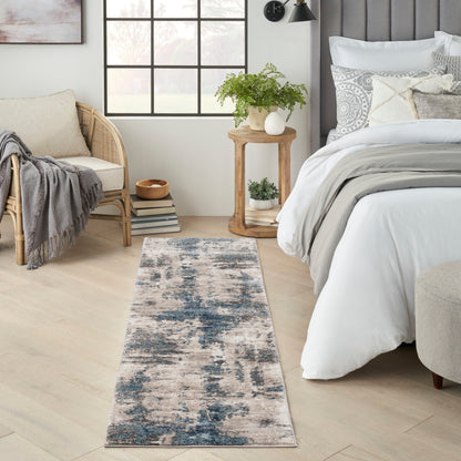 Abstract blue and beige runner rug along a light wood floor beside a white bed and rattan chair in a bright bedroom.