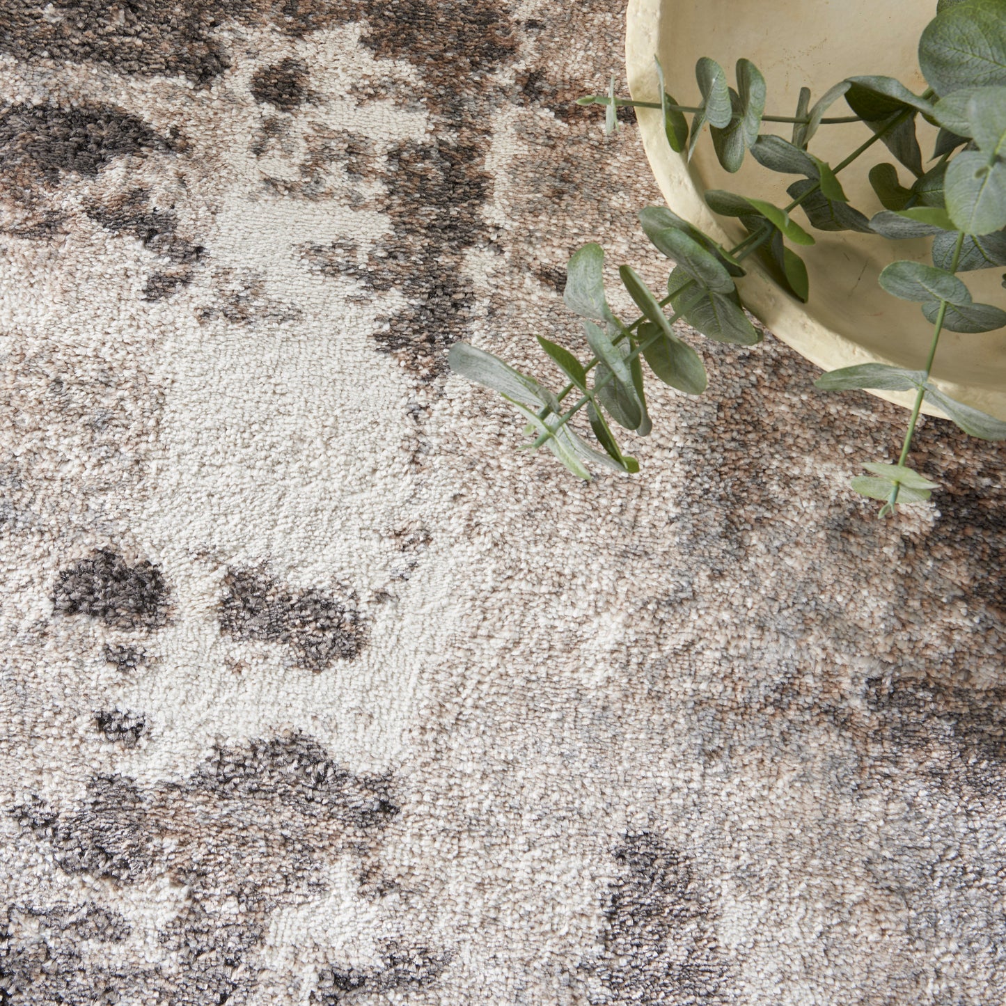 Beige textured rug with brown and gray splotches; eucalyptus plant in a beige pot overlaps the rug edge.