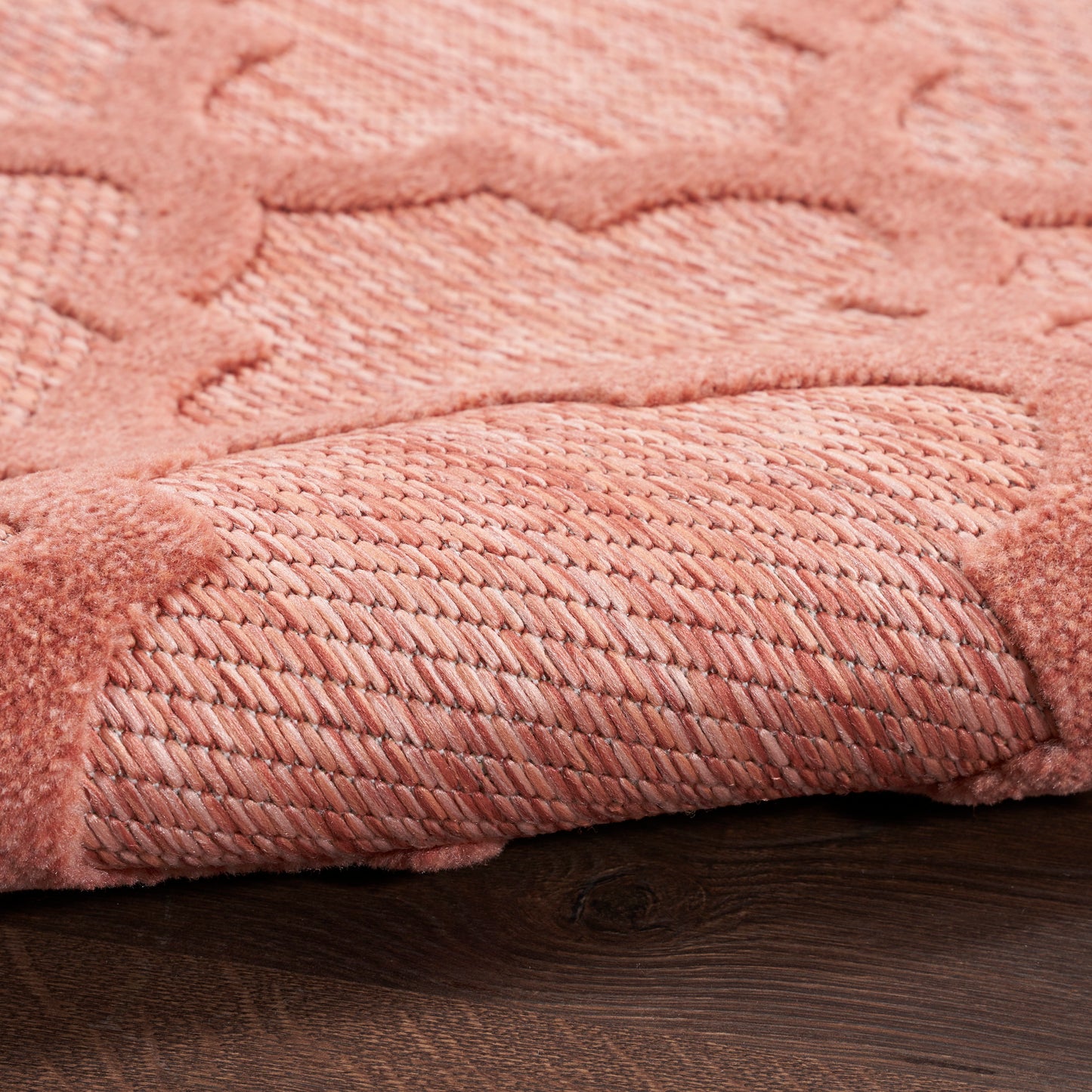 Dusty rose pink textured area rug with damask pattern; rolled edge reveals woven underside on dark wood floor.