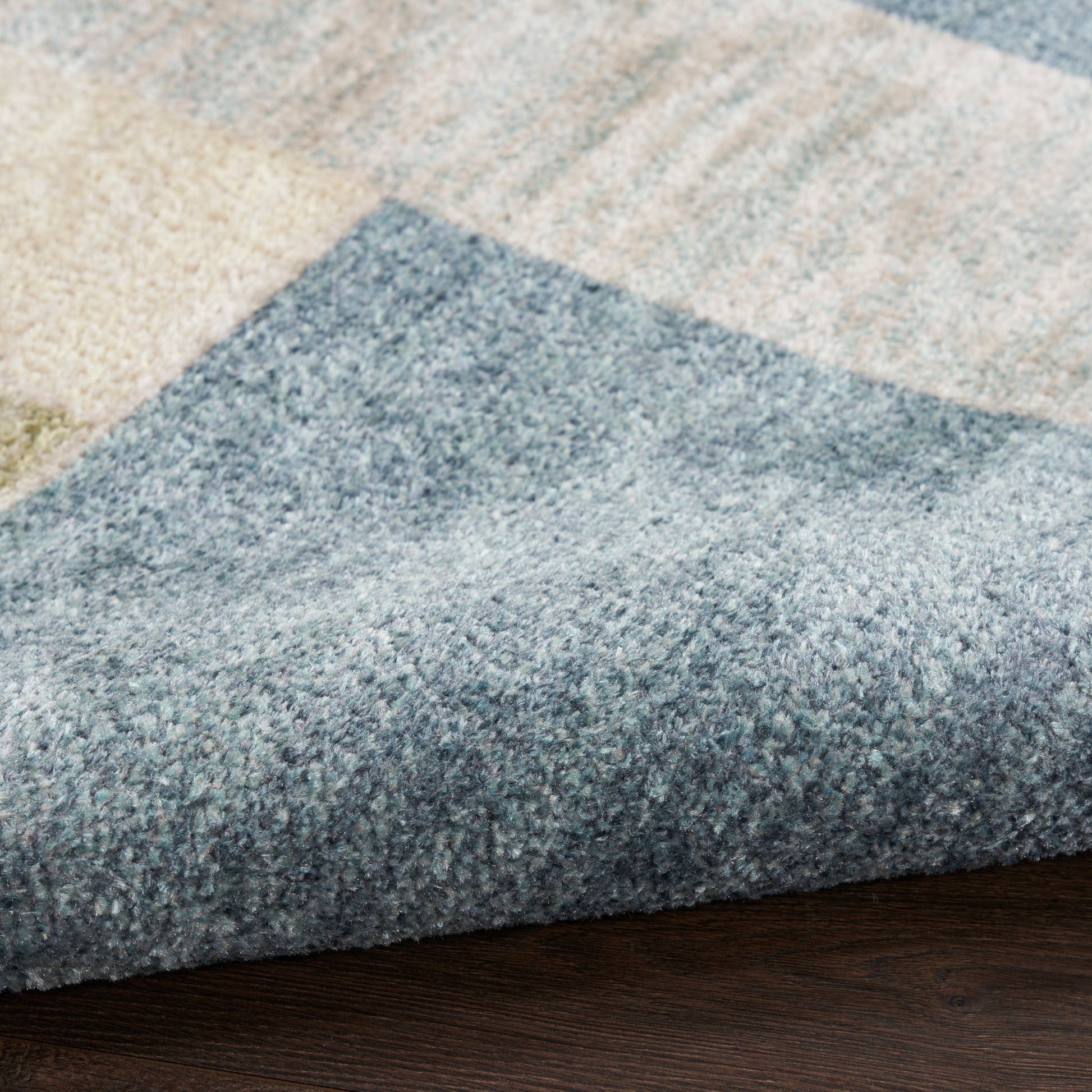 Blue-gray and beige geometric area rug with plush pile, folded edge on dark wood floor.