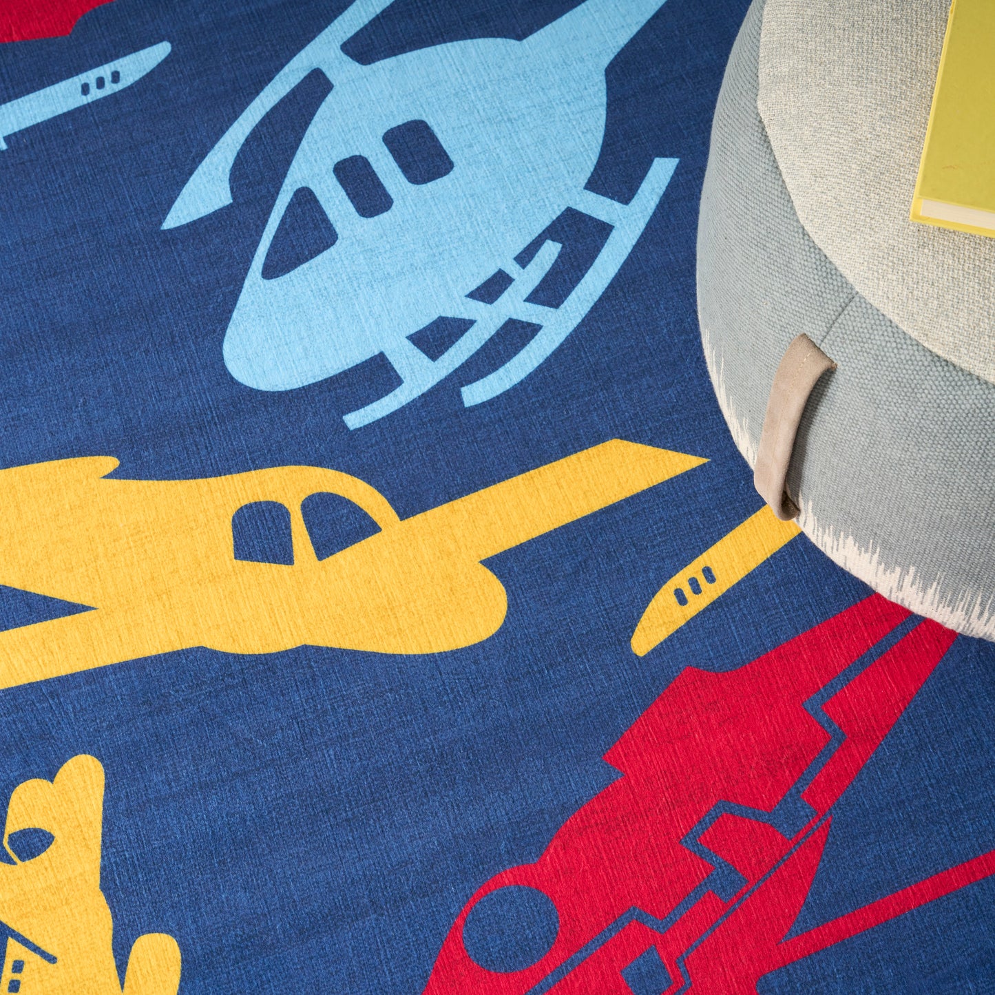 Blue kids' rug with yellow and light-blue airplane silhouettes; gray round pouf with tan strap nearby.