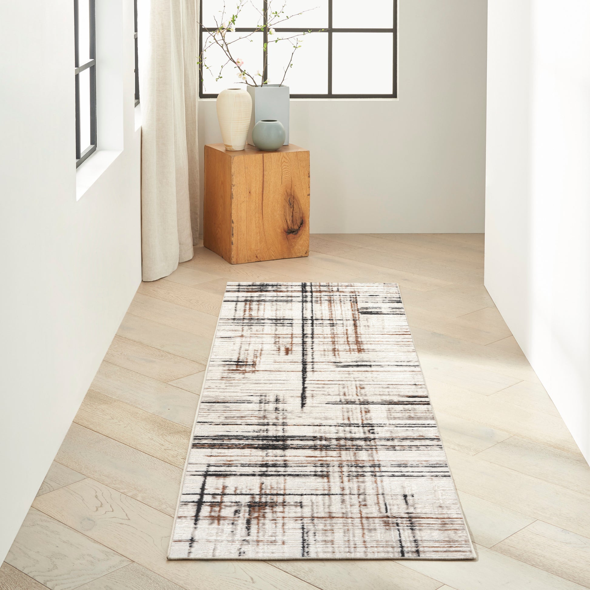 Long rectangular runner rug with white base & black-brown abstract grid in a bright hallway; beside a wooden block table with vases.