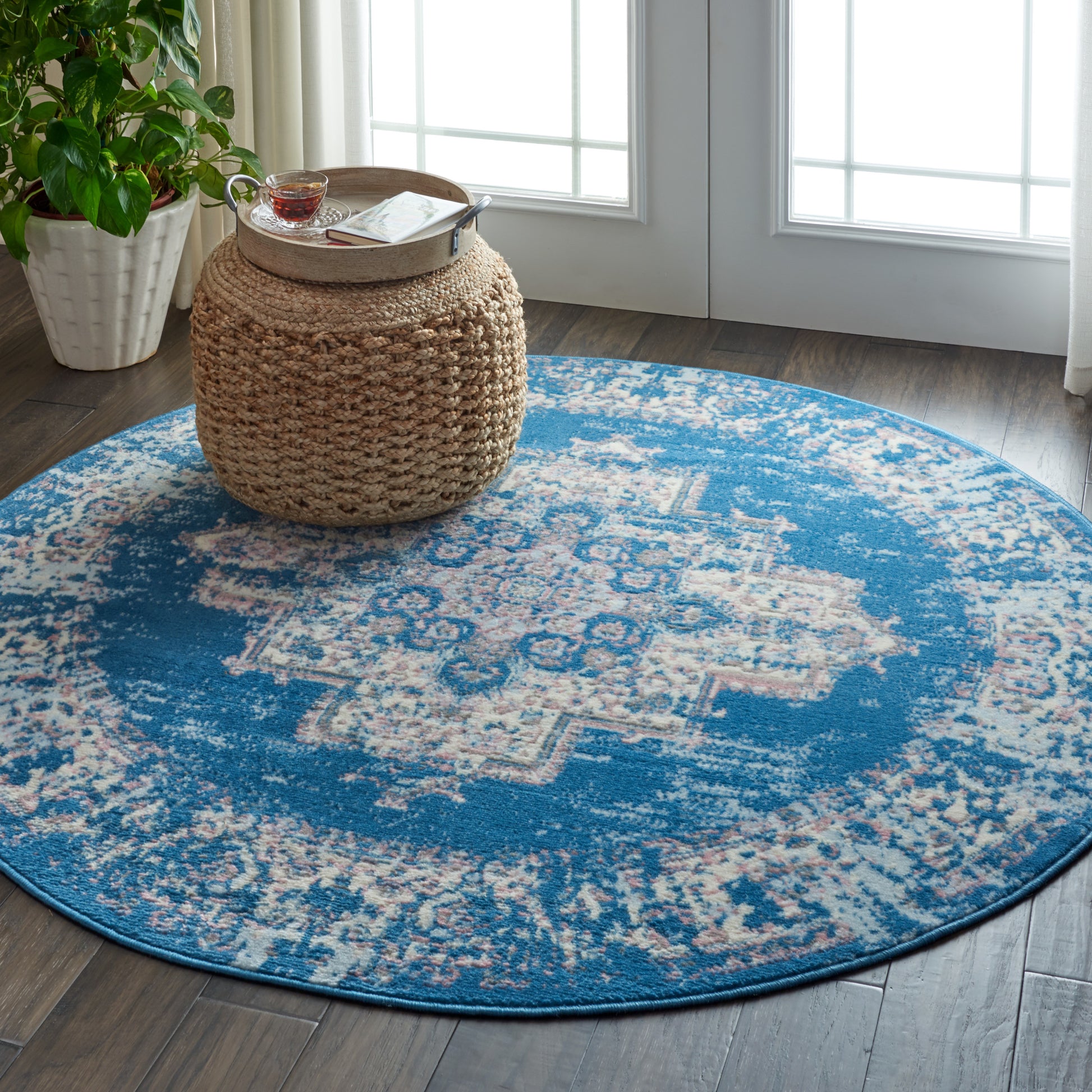 Round blue and cream patterned rug on wood floor with a natural woven pouf and potted plant near glass doors.