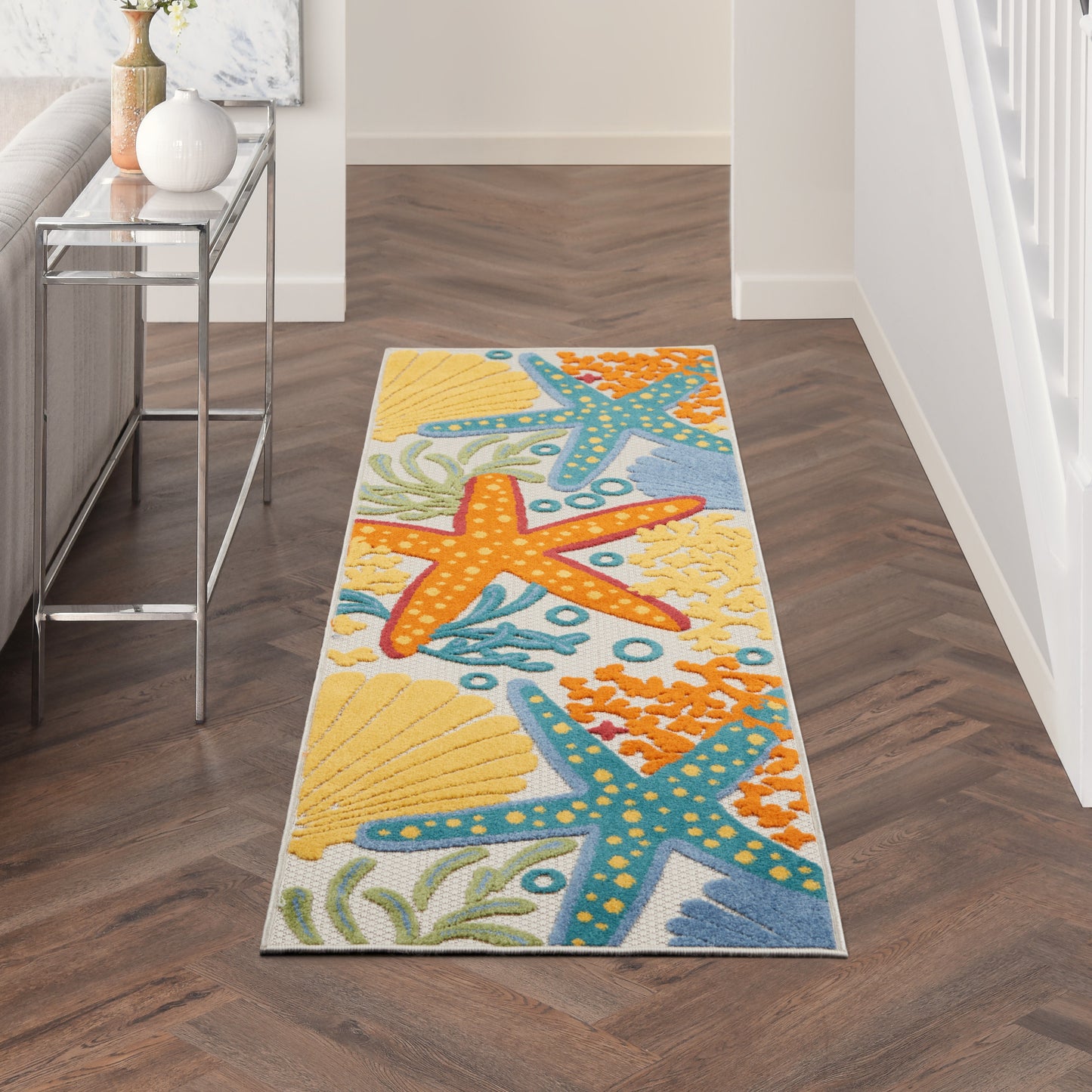 Coastal rectangular runner rug with orange blue and yellow starfish and seashell patterns in a hallway near a glass console