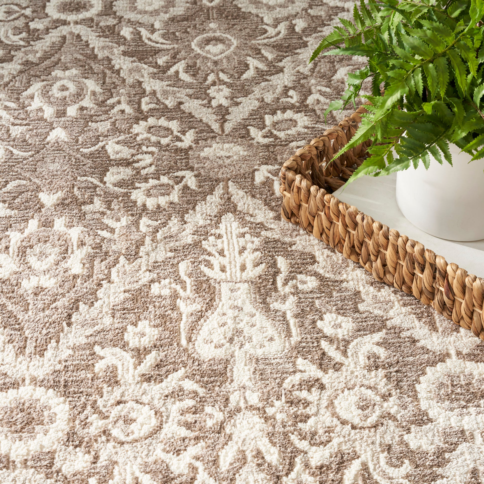 Beige-brown oriental rug with floral motif; woven natural basket on white tray holding a green fern.