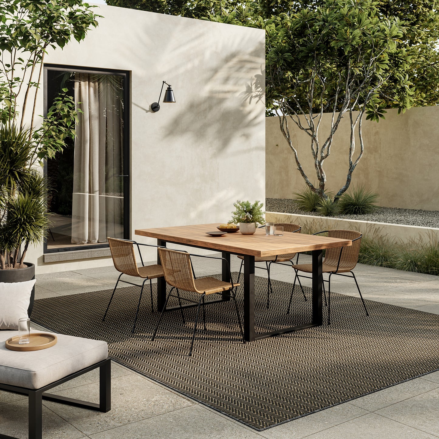 Outdoor patio scene featuring a modern 6' x 9' rug beneath a rectangular wooden dining table, surrounded by woven rattan chairs and greenery.
