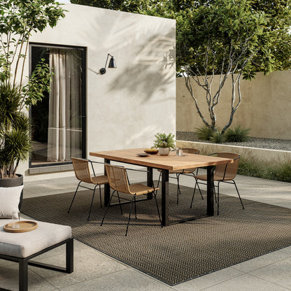Outdoor patio scene featuring a modern 6' x 9' rug beneath a rectangular wooden dining table, surrounded by woven rattan chairs and greenery.