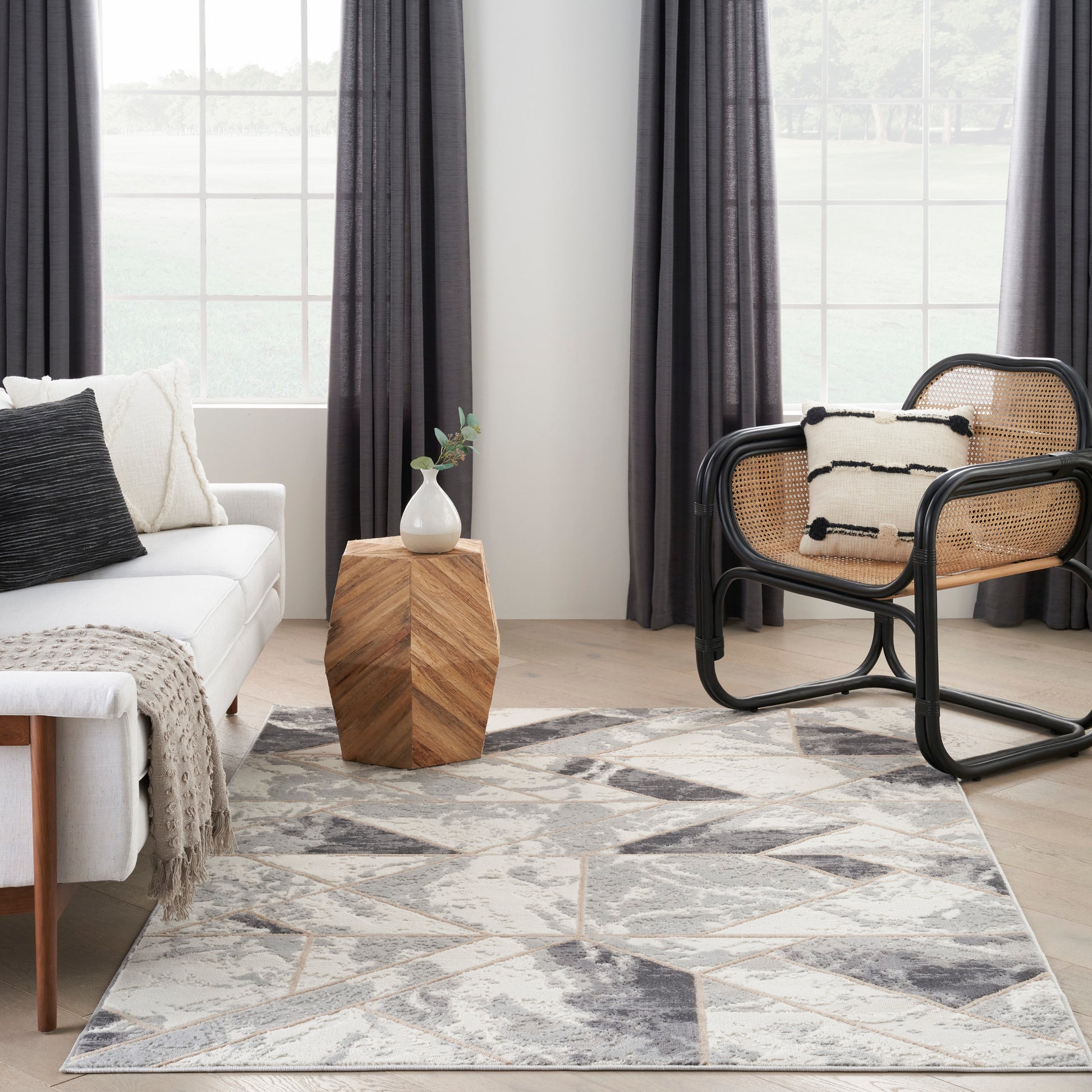 Modern living room: white sofa, black-rattan chair, geometric grey rug, wooden hexagonal side table with vase.