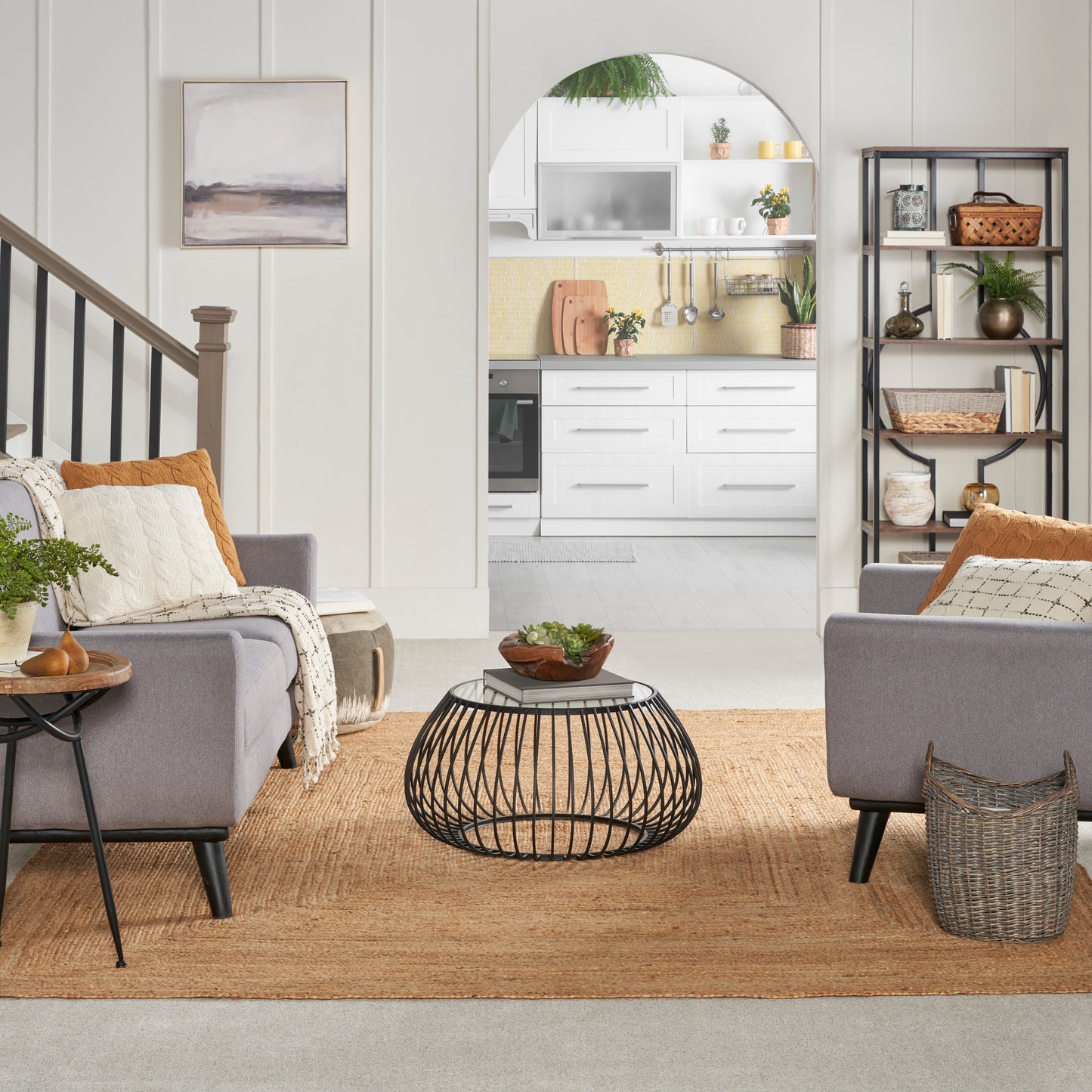 Living room with gray upholstered chairs, round black wire coffee table, natural jute rug, arch to white kitchen.