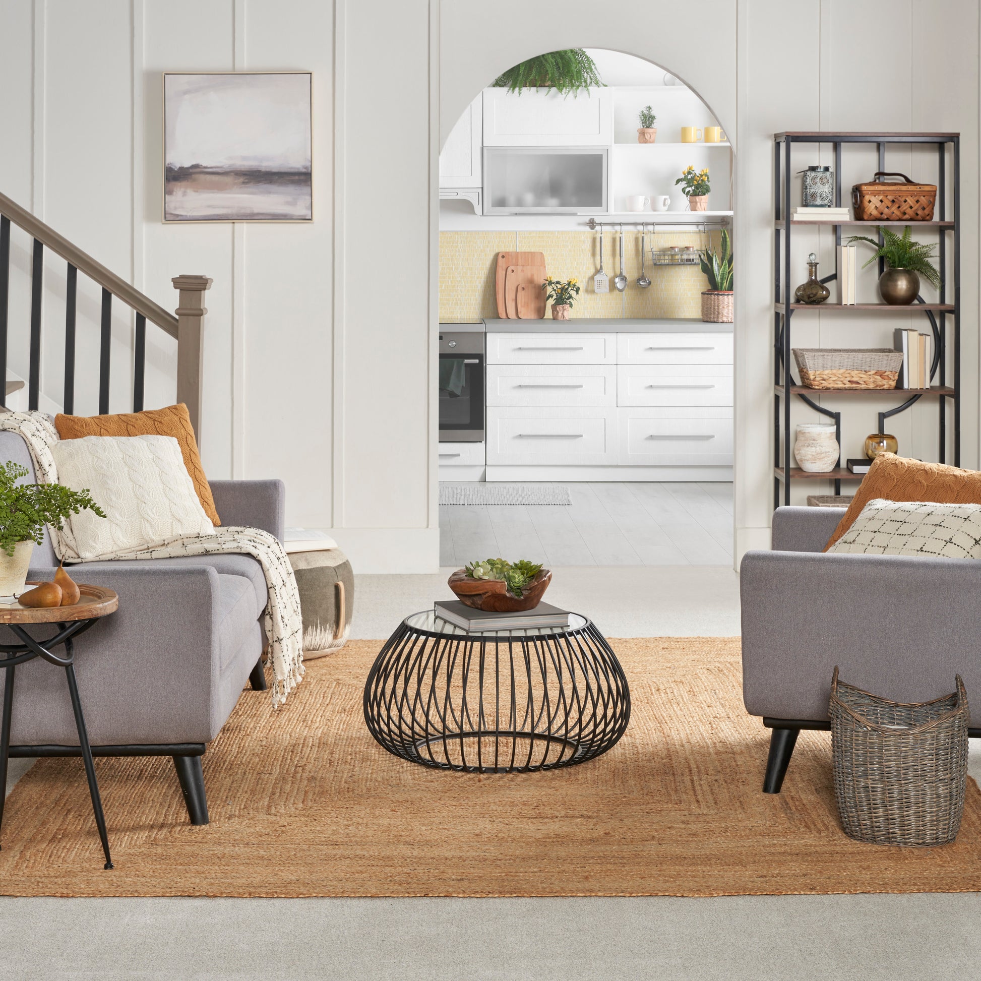 Living room with gray upholstered chairs, round black wire coffee table, natural jute rug, arch to white kitchen.