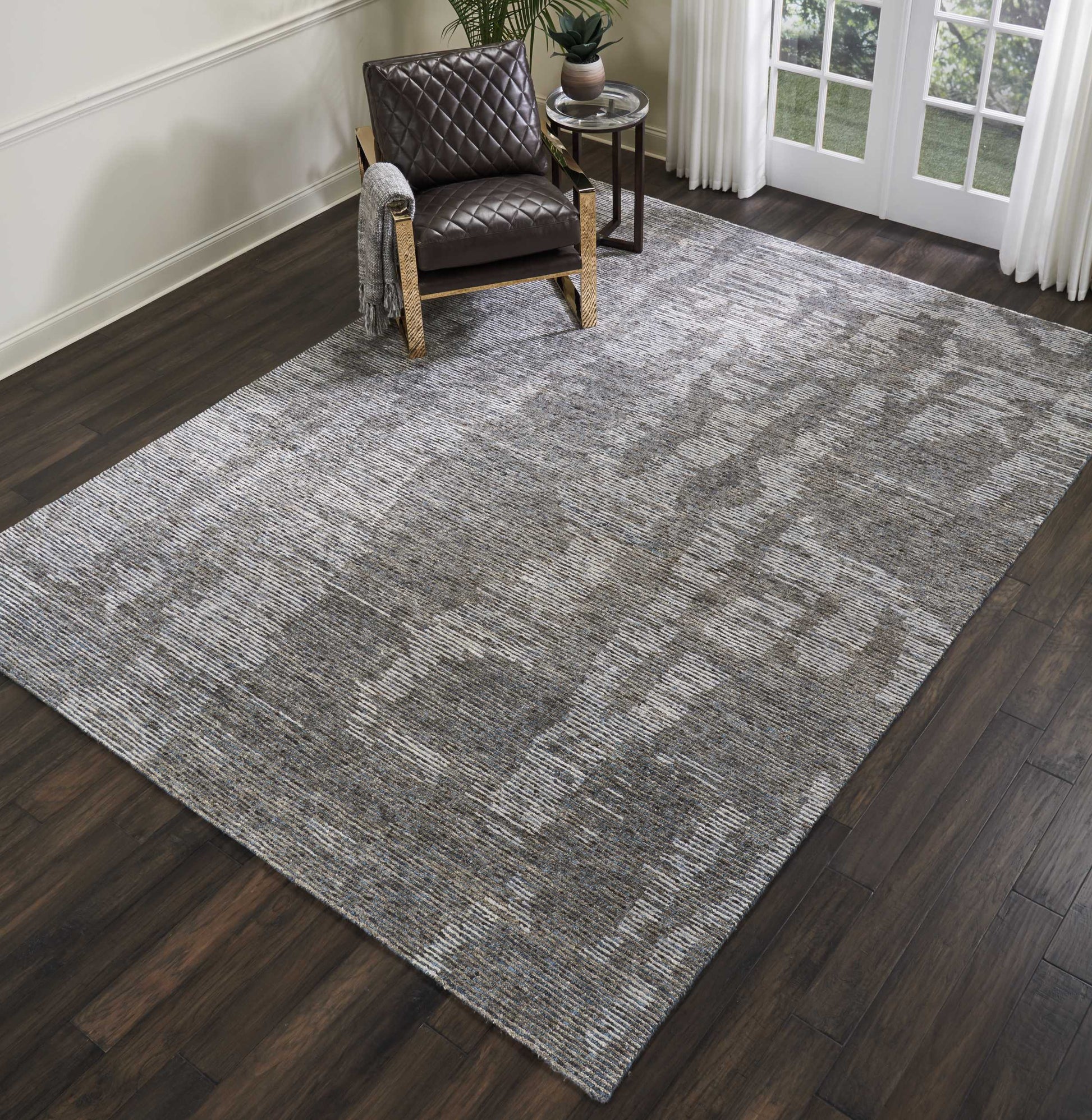 Large gray textured area rug on dark wood floor with a brown quilted armchair, a small side table, plant, and curtains.