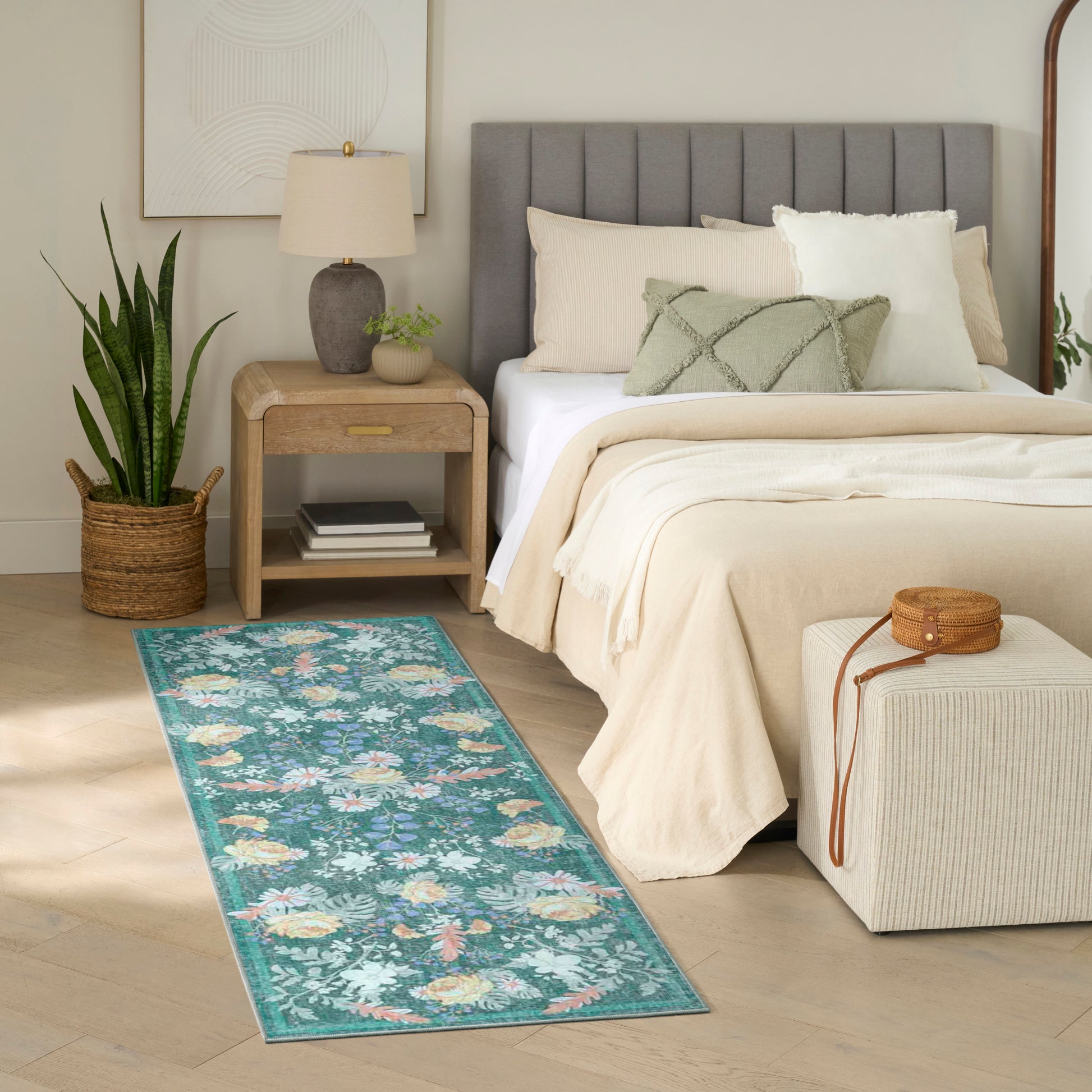 Teal floral runner rug beside beige bed with gray upholstered headboard, wooden nightstand, potted plant, and woven ottoman.
