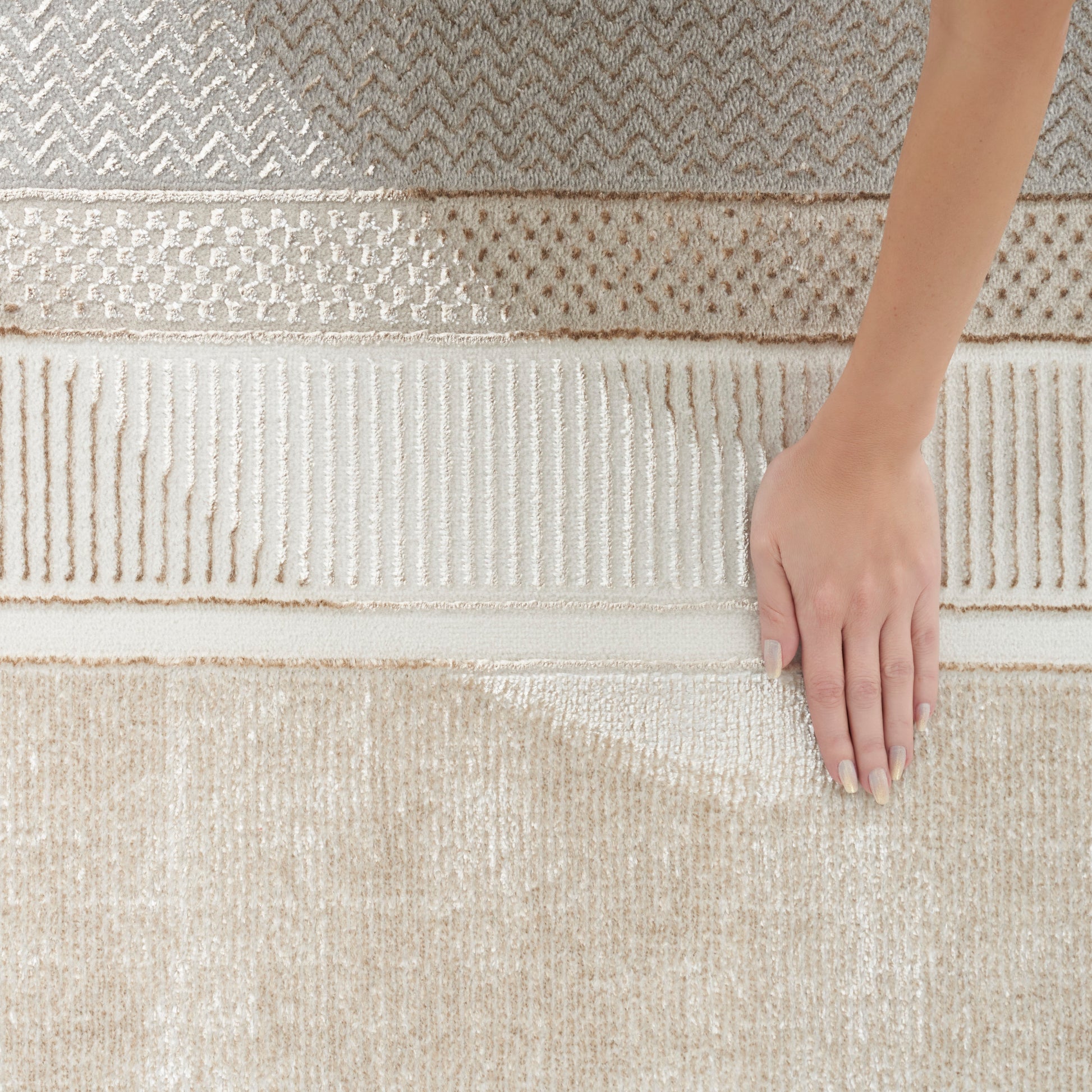 Cream-beige layered woven area rug with chevron, lattice and ribbed textures; hand resting on edge.