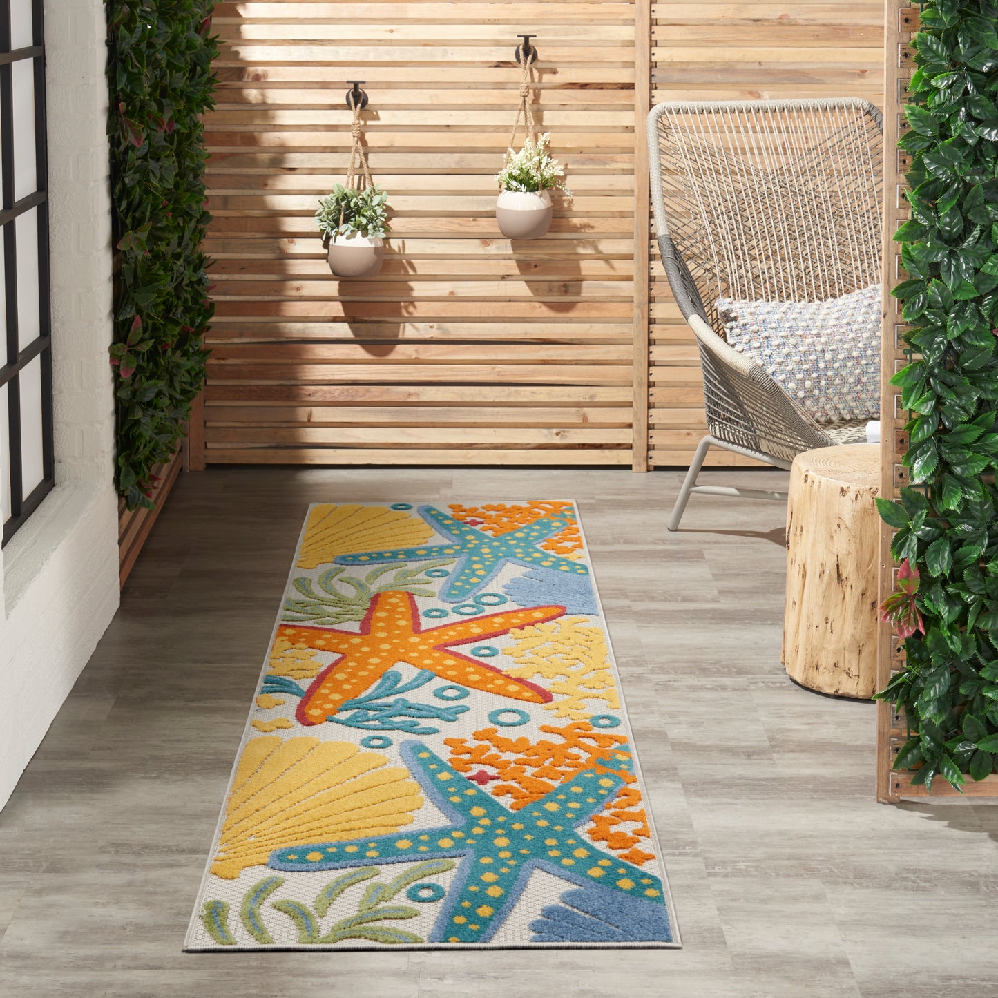 Coastal 6' runner rug with starfish and seaweed in bright blues and yellows; outdoor patio scene with wood deck, wicker chair and hanging planters.
