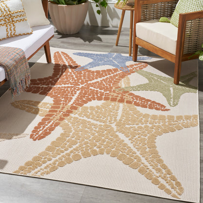 Outdoor rug with oversized starfish motifs in tan, orange, blue, and sage on a beige background beside wicker seating.