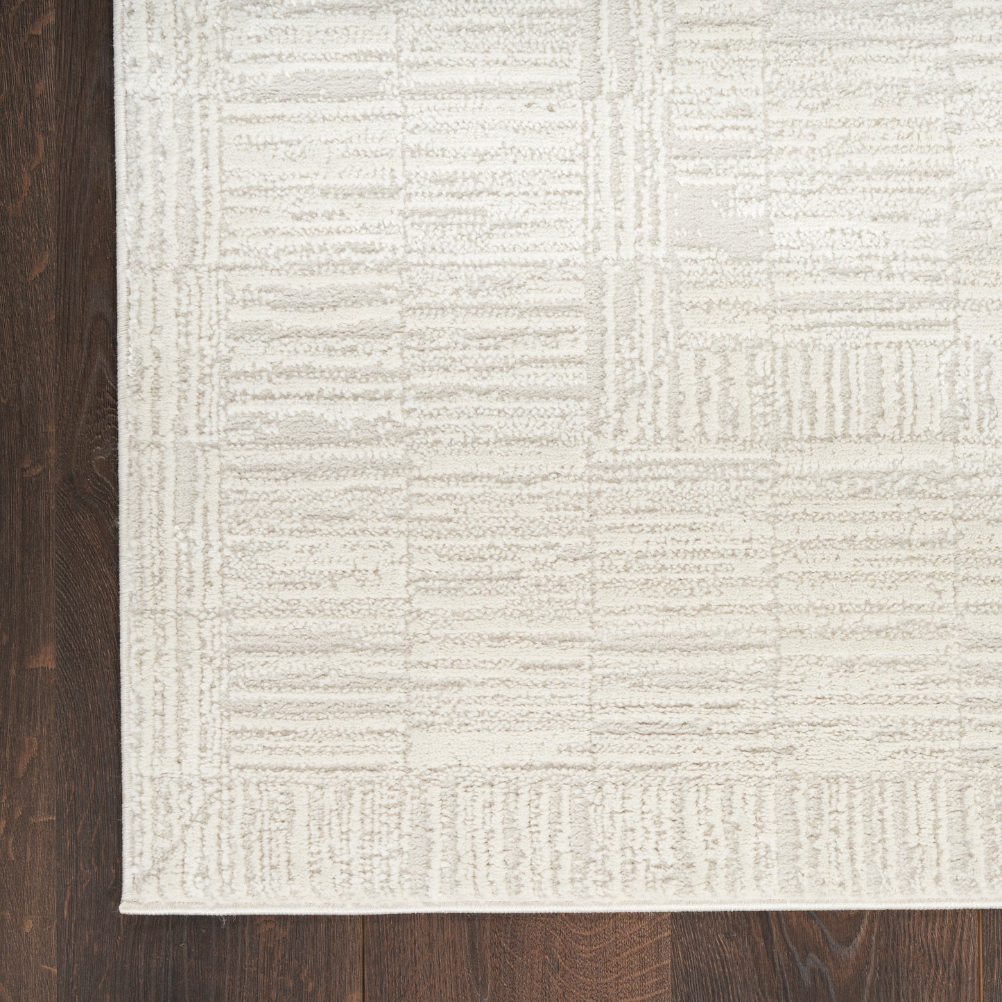 Beige textured rectangular area rug with geometric patchwork on dark wood floor; plush loop pile, contemporary style.
