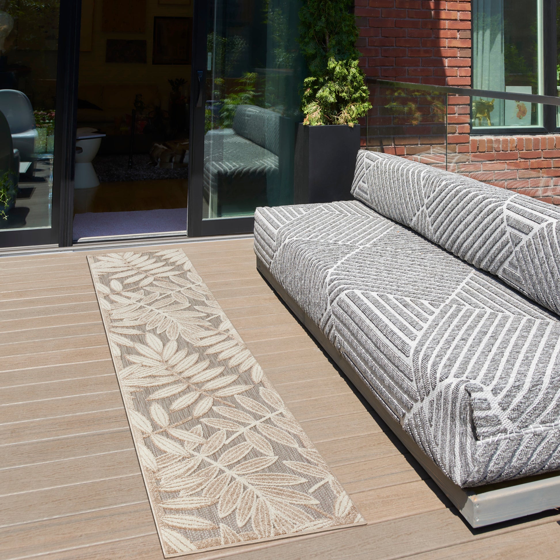 Outdoor wooden deck with a tropical beige leaf-pattern 10' runner rug beside glass doors and a brick wall.