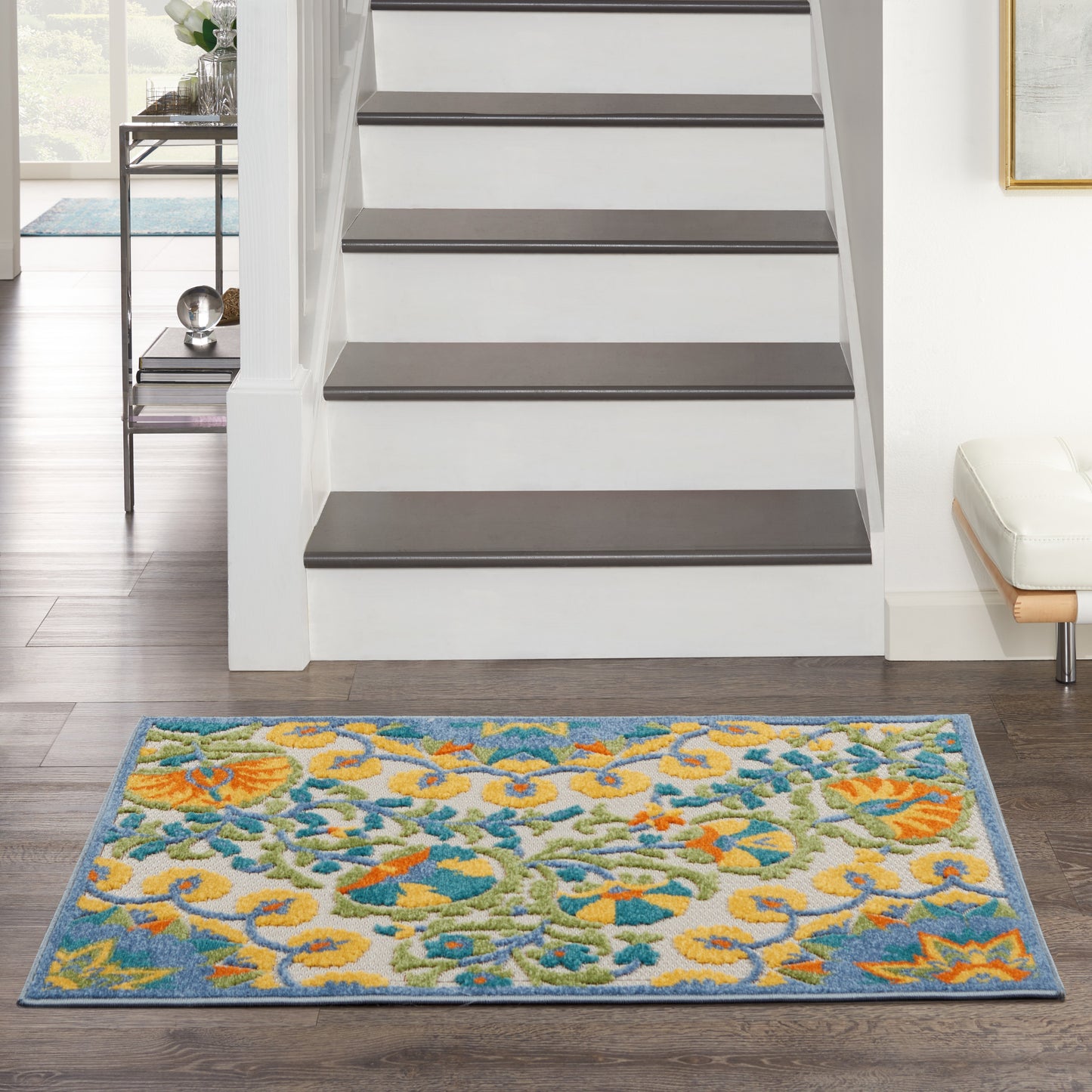 Vibrant blue-bordered floral area rug with yellow, teal and green flowers on dark wood floor at the foot of white stairs.