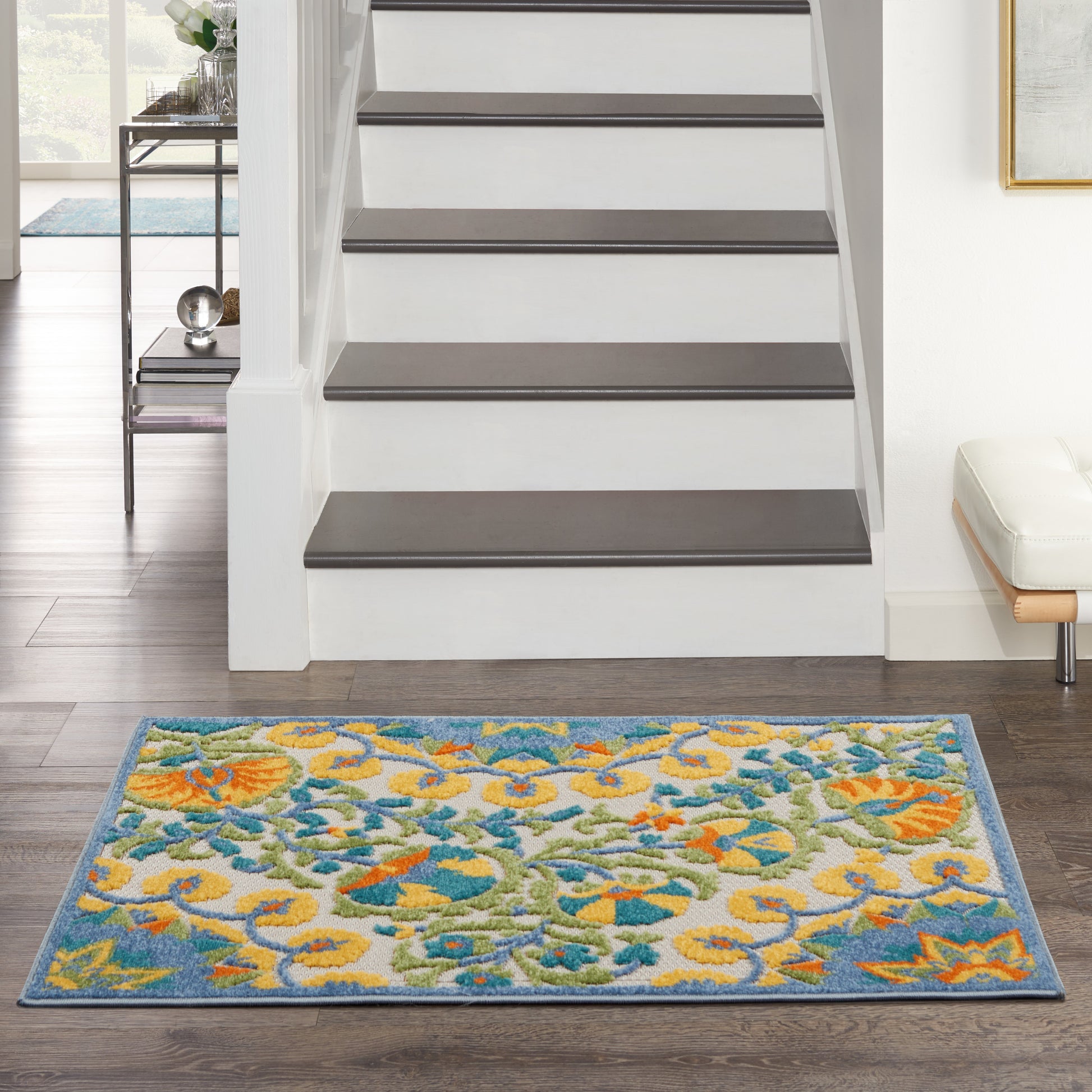 Vibrant blue-bordered floral area rug with yellow, teal and green flowers on dark wood floor at the foot of white stairs.