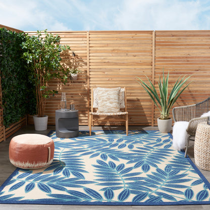 Outdoor patio scene featuring blue palm-leaf rug, wicker seating, wooden slat fence, potted plants and orange pouf.