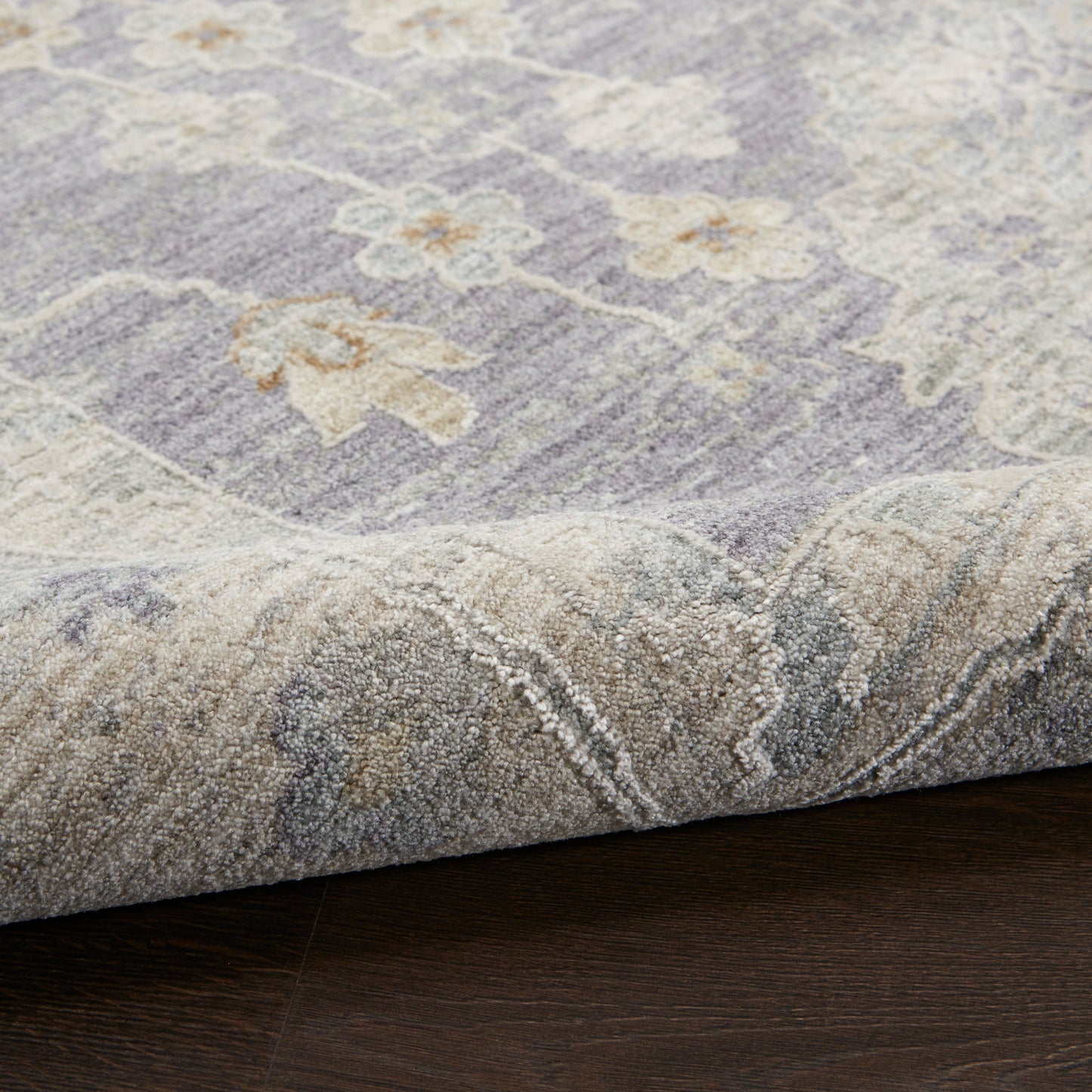 Lavender-gray floral rug with cream blossoms, plush pile, folded edge on dark wood floor.