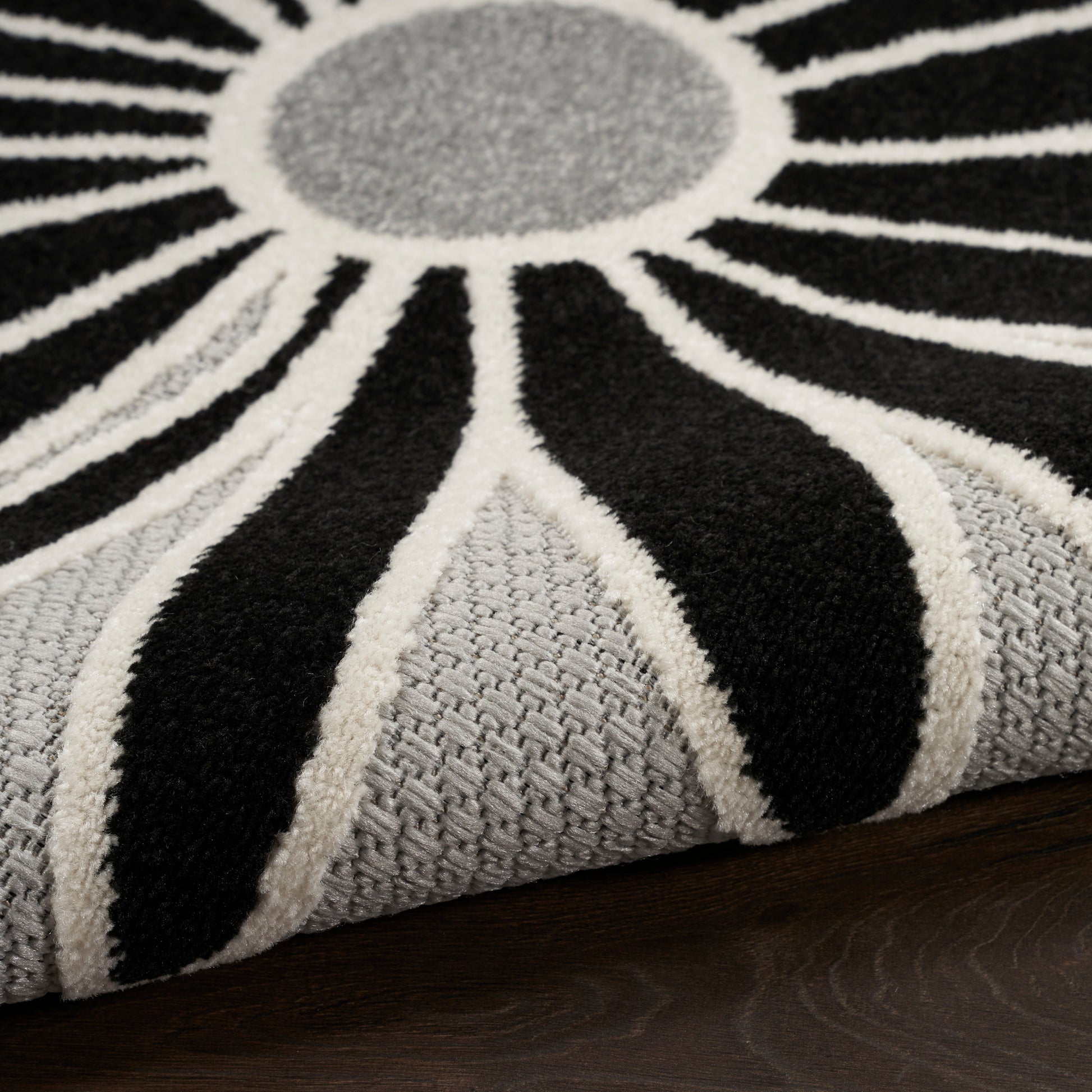 Close-up of a gray and black sunburst rug with white radiating lines and a textured gray base on dark wood.
