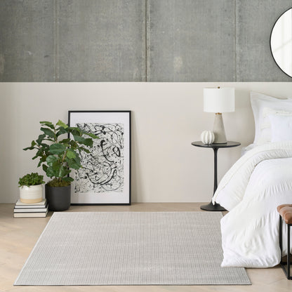 Modern bedroom scene with white bedding, light gray rug, black-framed abstract art, potted plant, books, and a black side table with a white lamp.