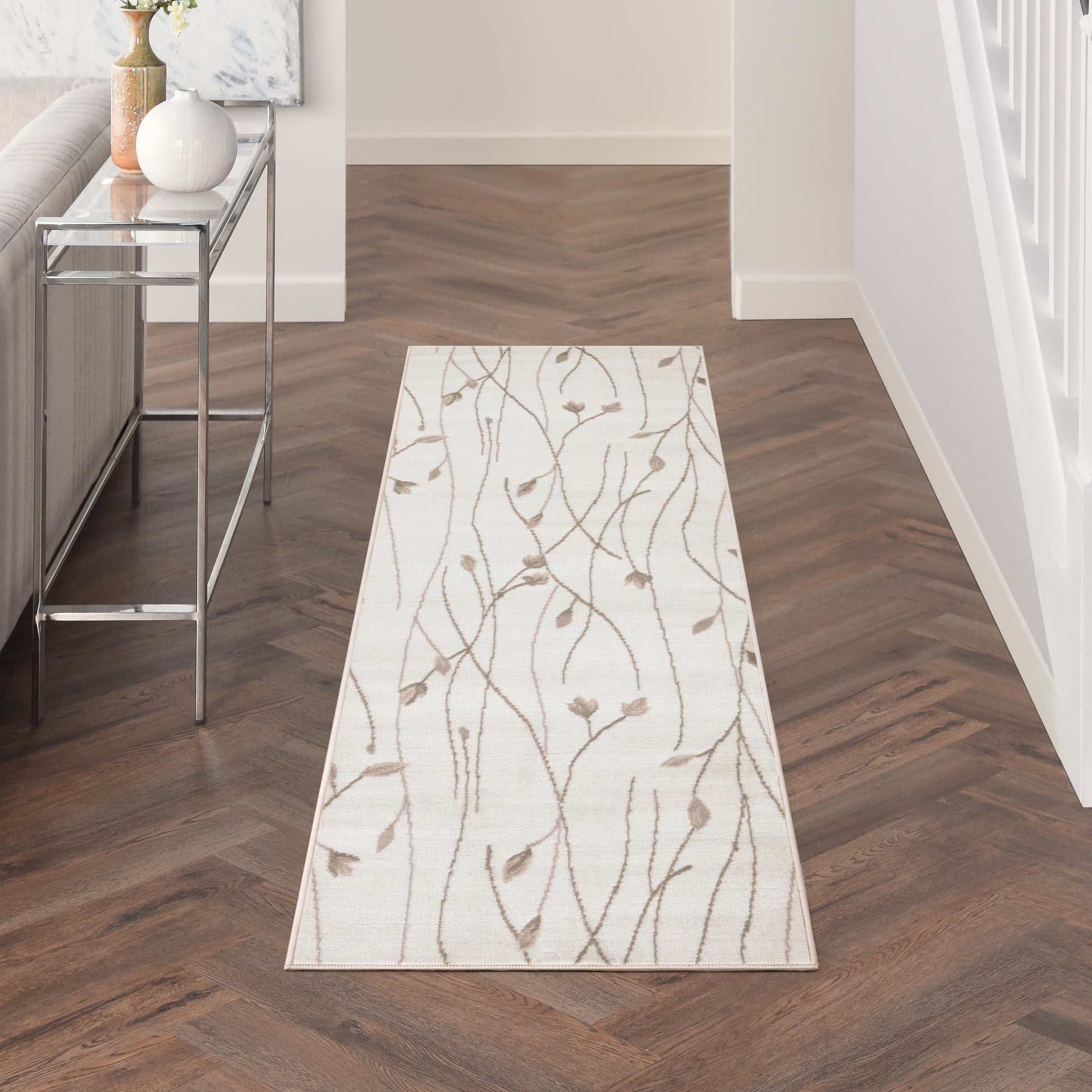 Beige hallway runner with brown branch-pattern rug on dark wood herringbone floor; chrome console table with vases nearby.