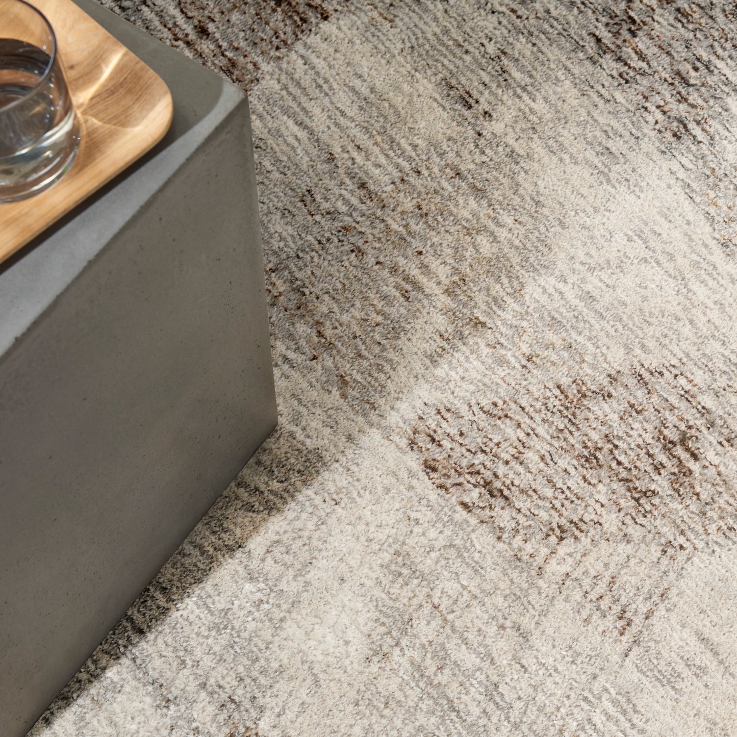 Gray concrete side table with a light wooden tray holding a glass of water, set on a beige-brown patterned rug.