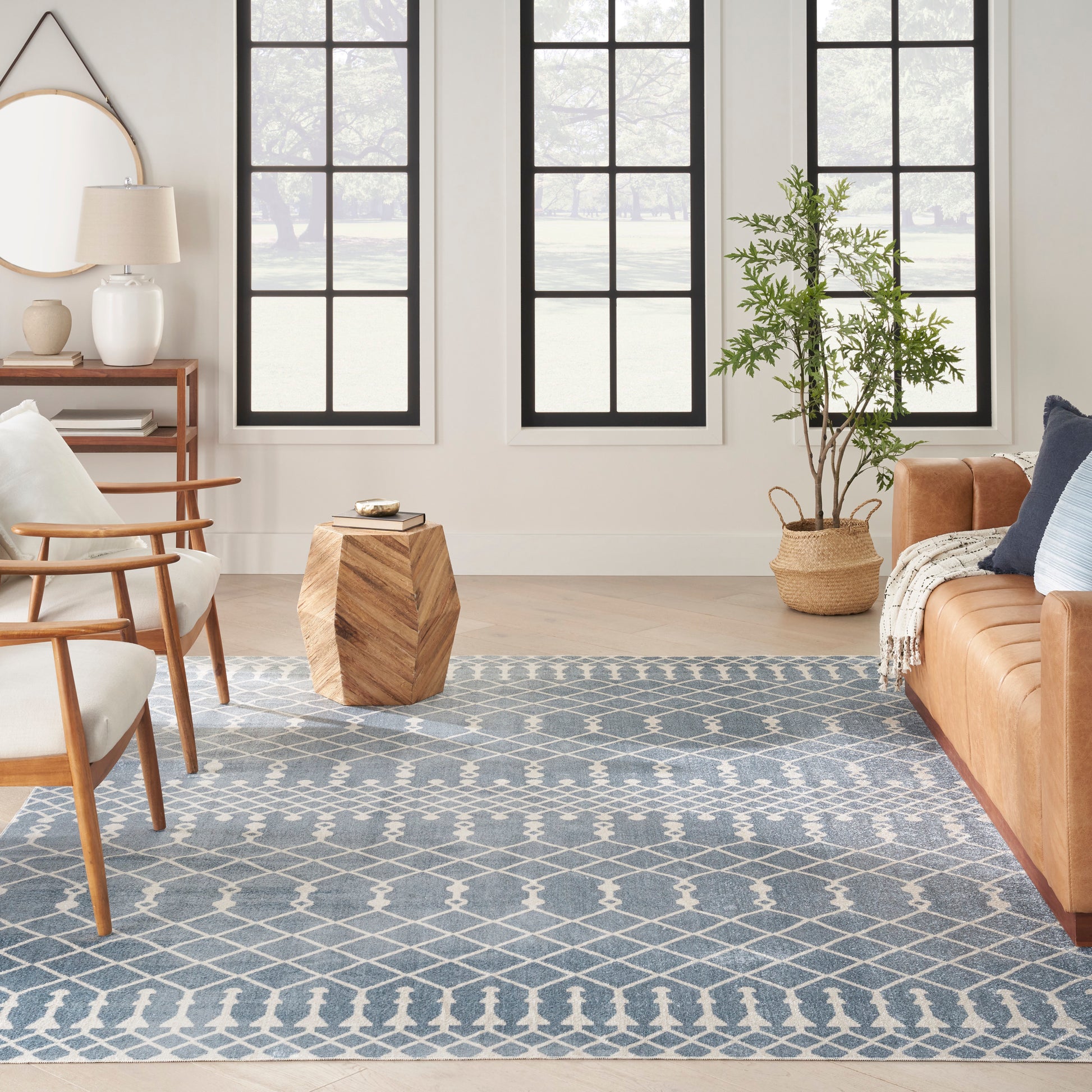 Bright living room with blue geometric rug, mid-century wooden chairs, tan leather sofa, woven plant basket, grid windows.