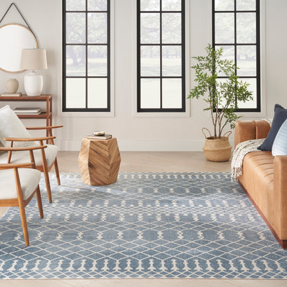 Bright living room with blue geometric rug, mid-century wooden chairs, tan leather sofa, woven plant basket, grid windows.