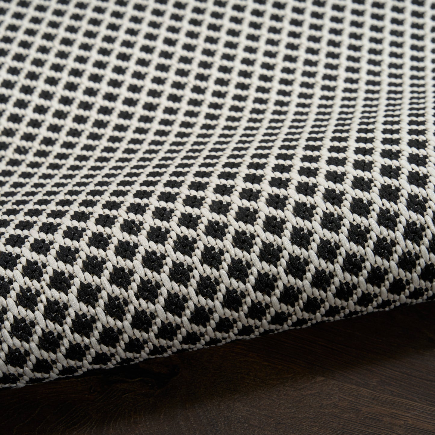 White and black woven rug with a small diamond lattice pattern, folded edge resting on dark wood surface.