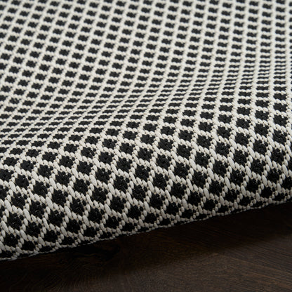 White and black woven rug with a small diamond lattice pattern, folded edge resting on dark wood surface.