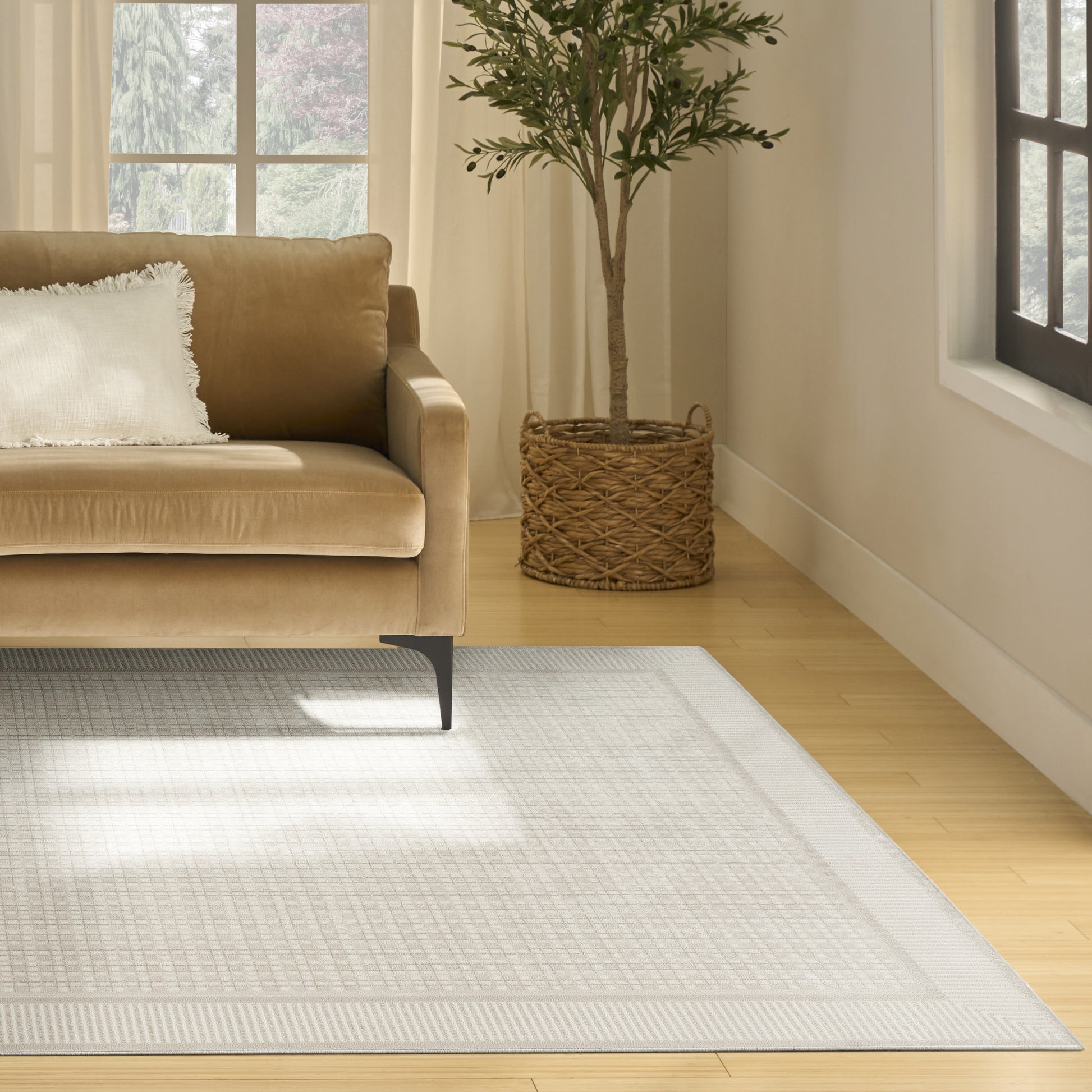 Geometric light gray area rug (8' x 10') in a sunlit living room with a beige velvet sofa, white fringe pillow, and a potted plant in a woven basket.
