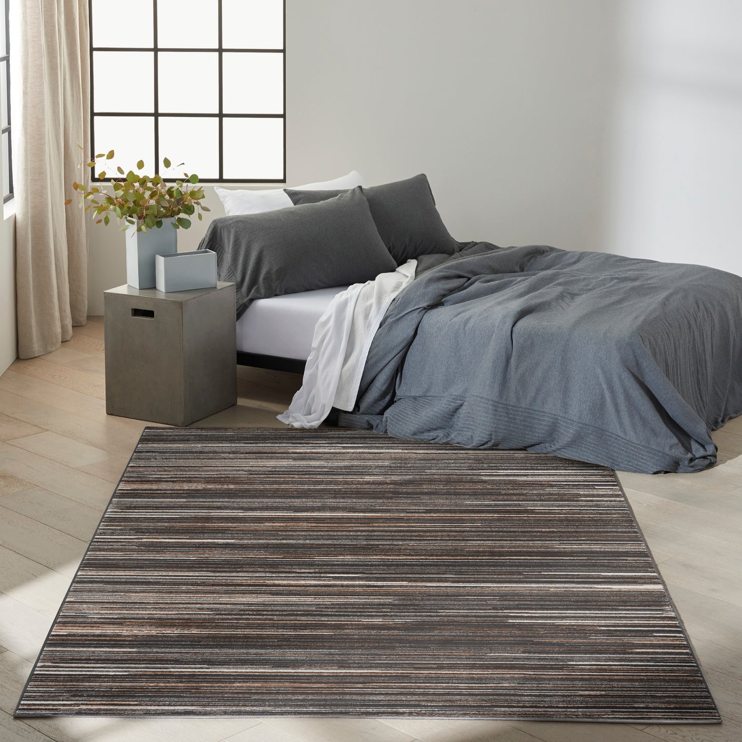 Modern bedroom with dark gray duvet, white sheet, gray pillows on bed, concrete nightstand with plant, brown striped rug.