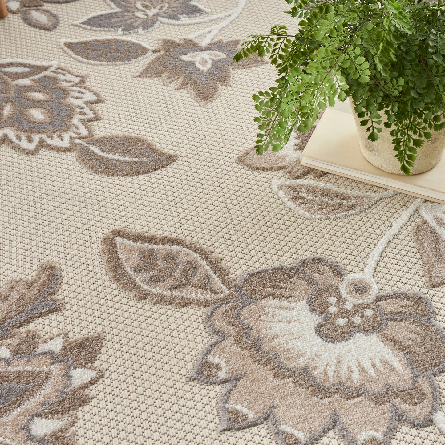 Beige floral-pattern area rug with brown and gray flowers; woven texture, with a green potted plant nearby.