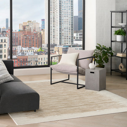 Modern living room with city skyline view, beige gradient rug, gray sectional, pink-beige metal-frame chair, concrete side table, and plant-filled shelving.