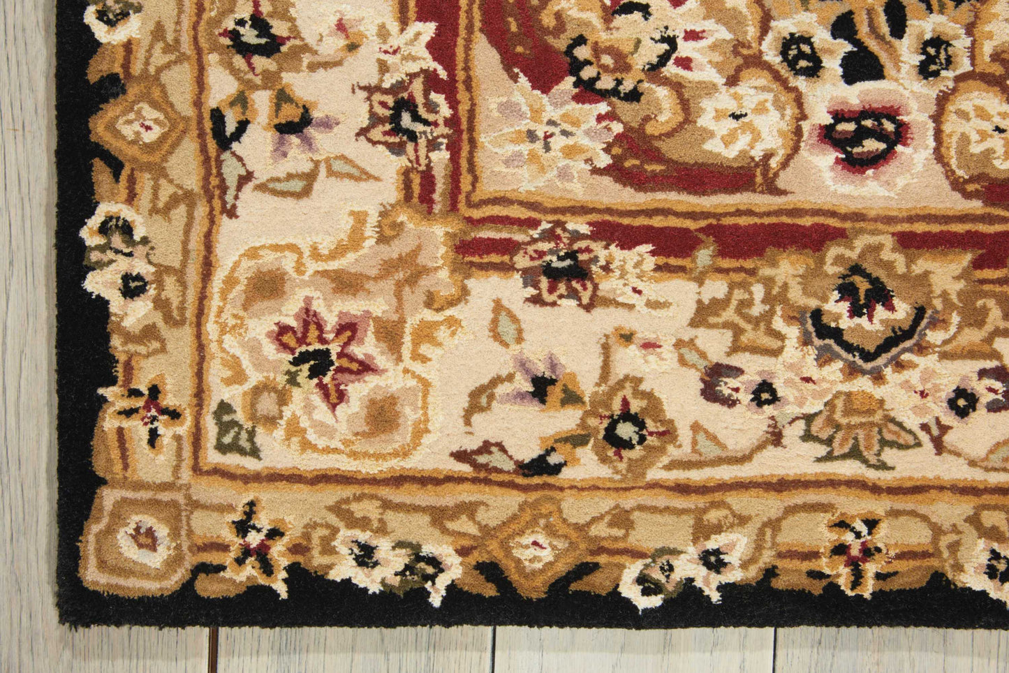 Corner of an ornate beige Oriental rug with cream, gold and red floral motifs and a thick black border on a wooden floor.