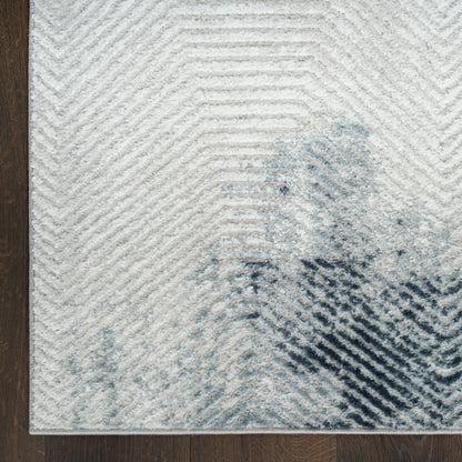 Light gray area rug with chevron texture and blue-black abstract blot near the corner on dark wood floor.