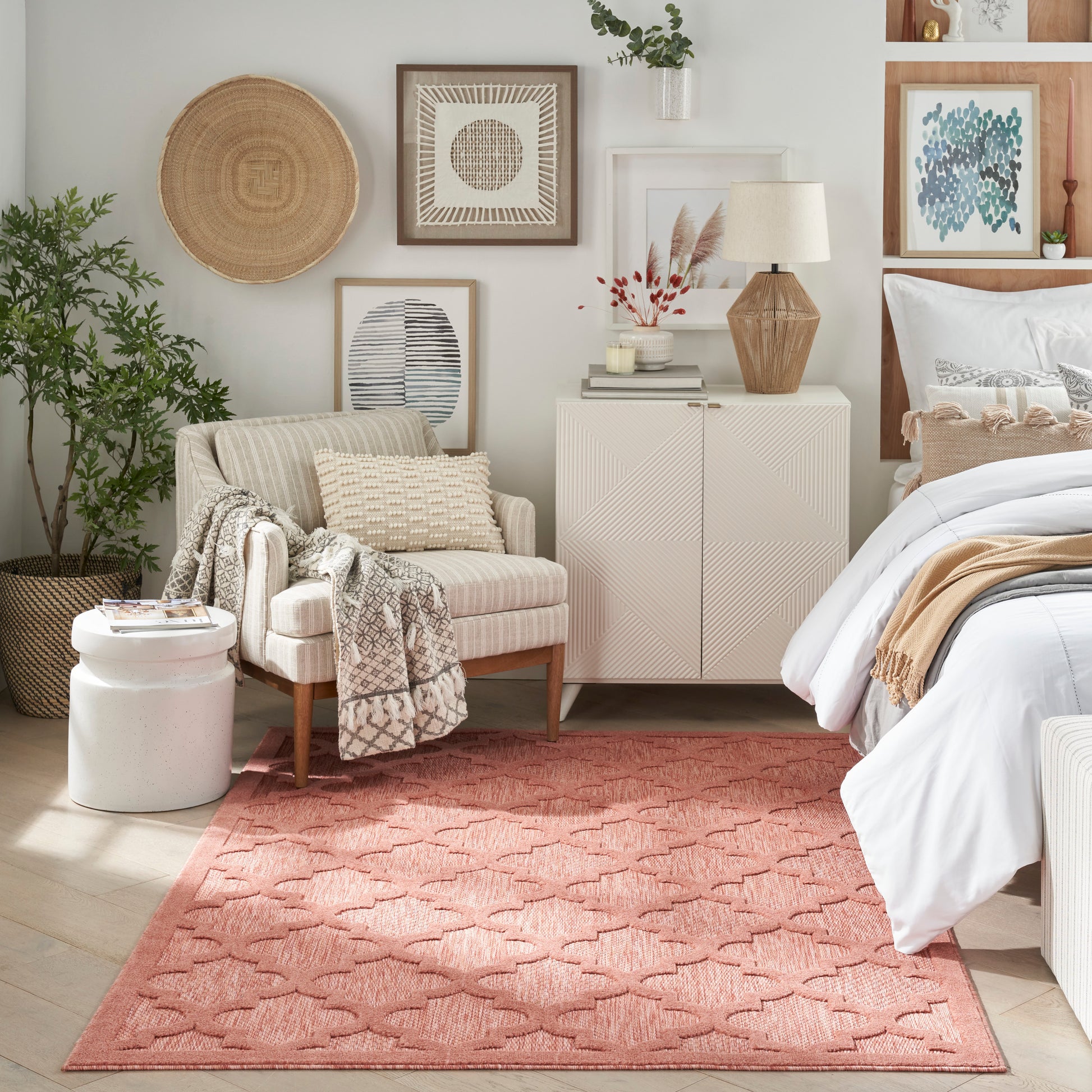 Cozy bedroom with pink geometric rug, beige armchair, white cabinet, bed with white linens, and framed wall art.