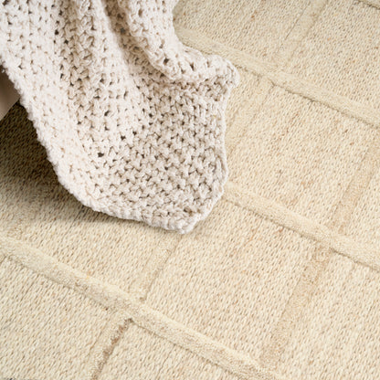 Cream chunky-knit throw draped over a beige textured rug with square block pattern.