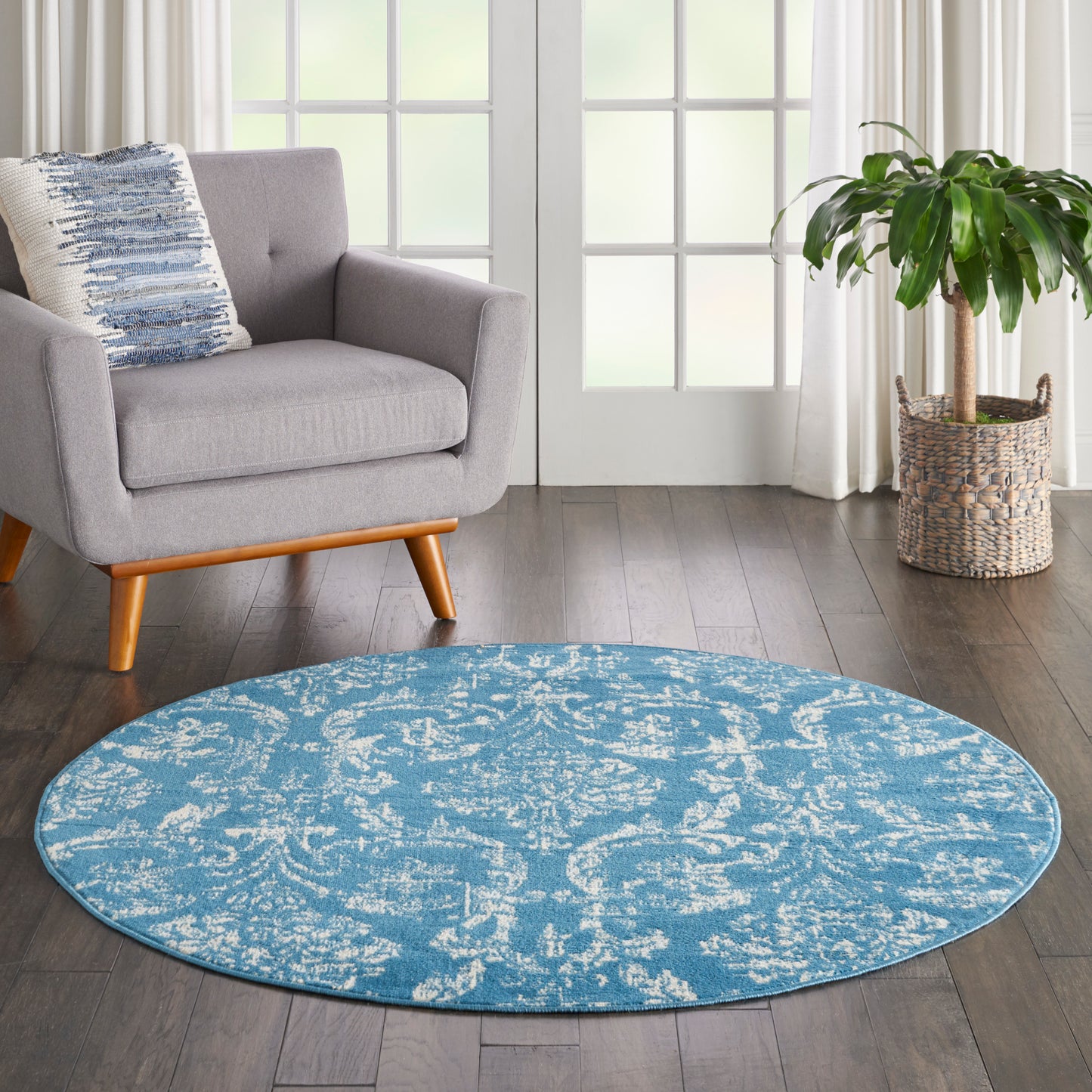 Round blue patterned rug on dark wood floor beside a gray mid-century armchair and a potted plant.