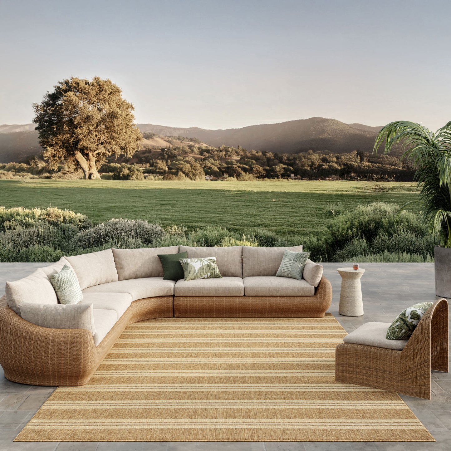 Curved tan wicker outdoor sectional with cream cushions and green pillows on striped jute rug; serene landscape beyond.
