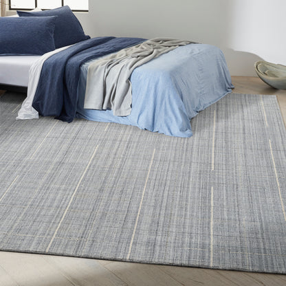 Bed with navy and light blue linens, gray throw, atop a large gray striped rug on light wood floor.