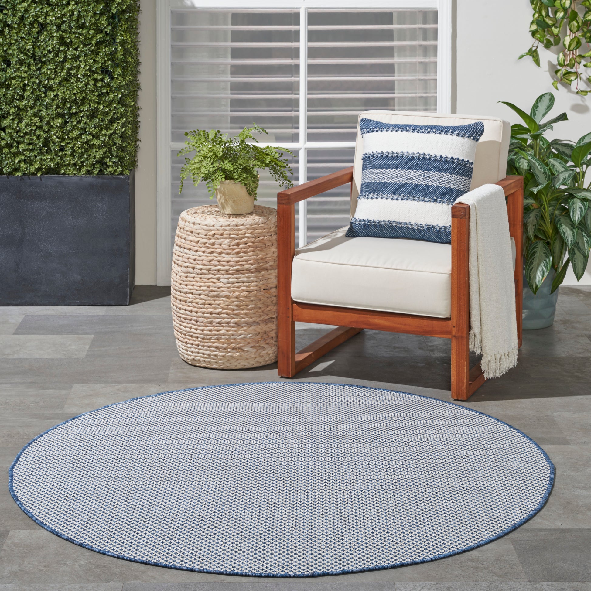 Round blue-and-white outdoor rug with wooden armchair, cream cushions, striped pillow, and woven side table with plant.