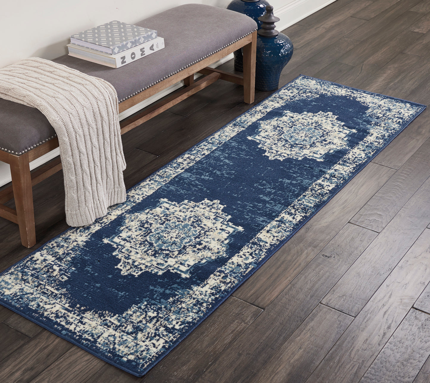 Navy blue distressed runner rug with ivory medallions on dark wood floor beside a gray padded bench and beige knit throw.